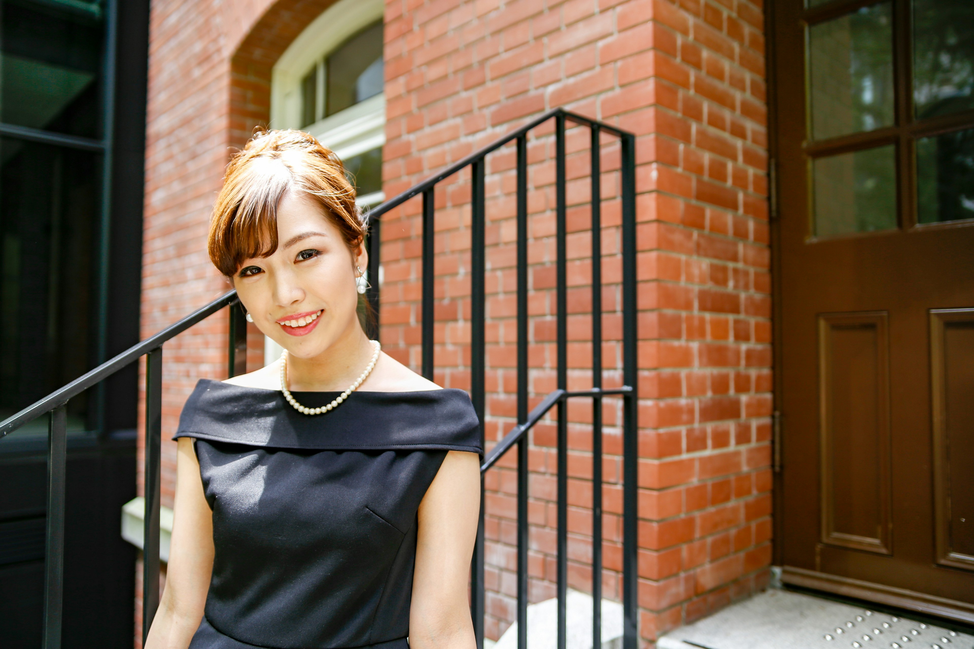 A woman in a black dress smiling while standing on stairs with a red brick wall in the background