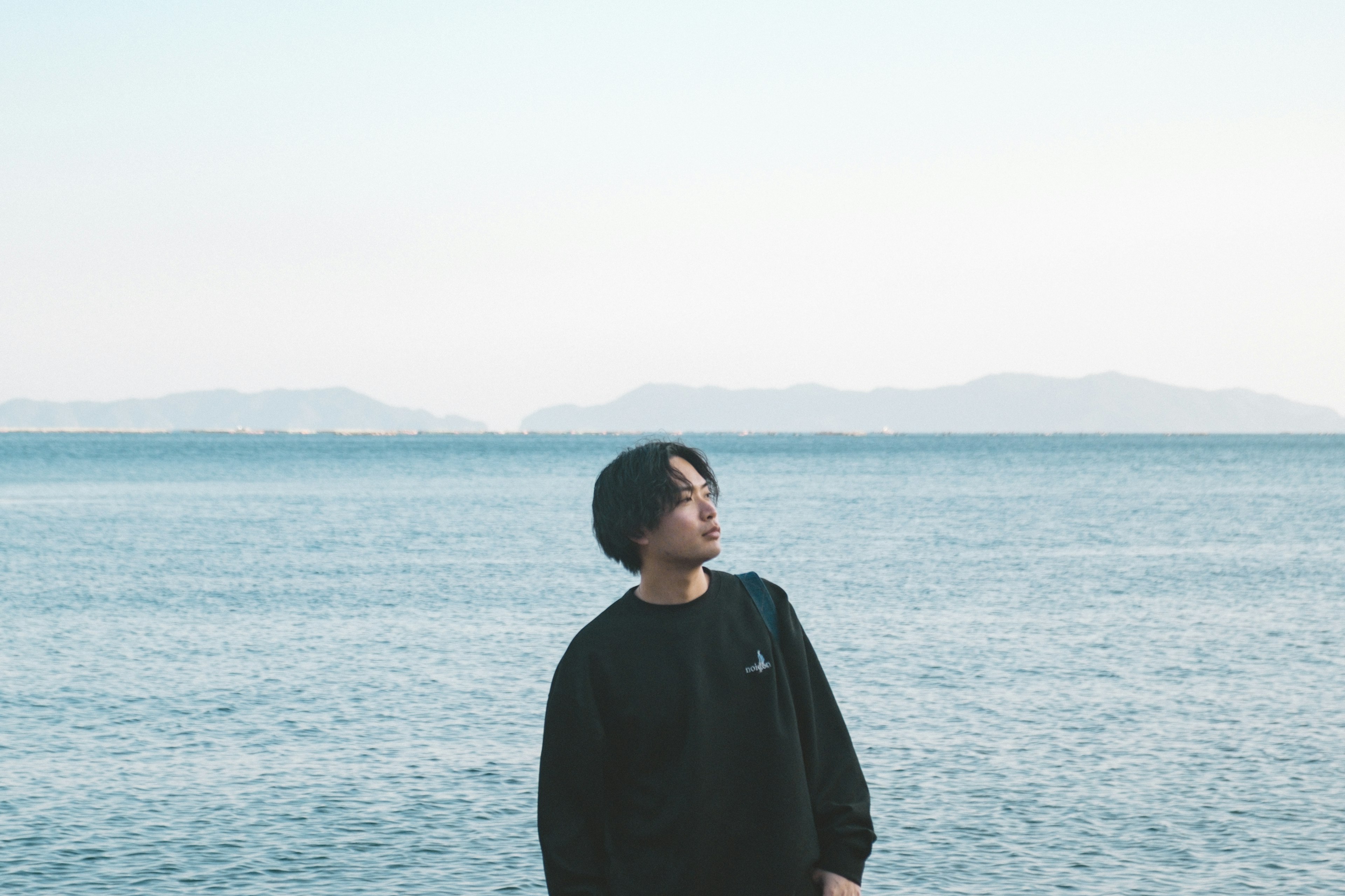 A young man standing by the seaside with a calm ocean and distant mountains in the background