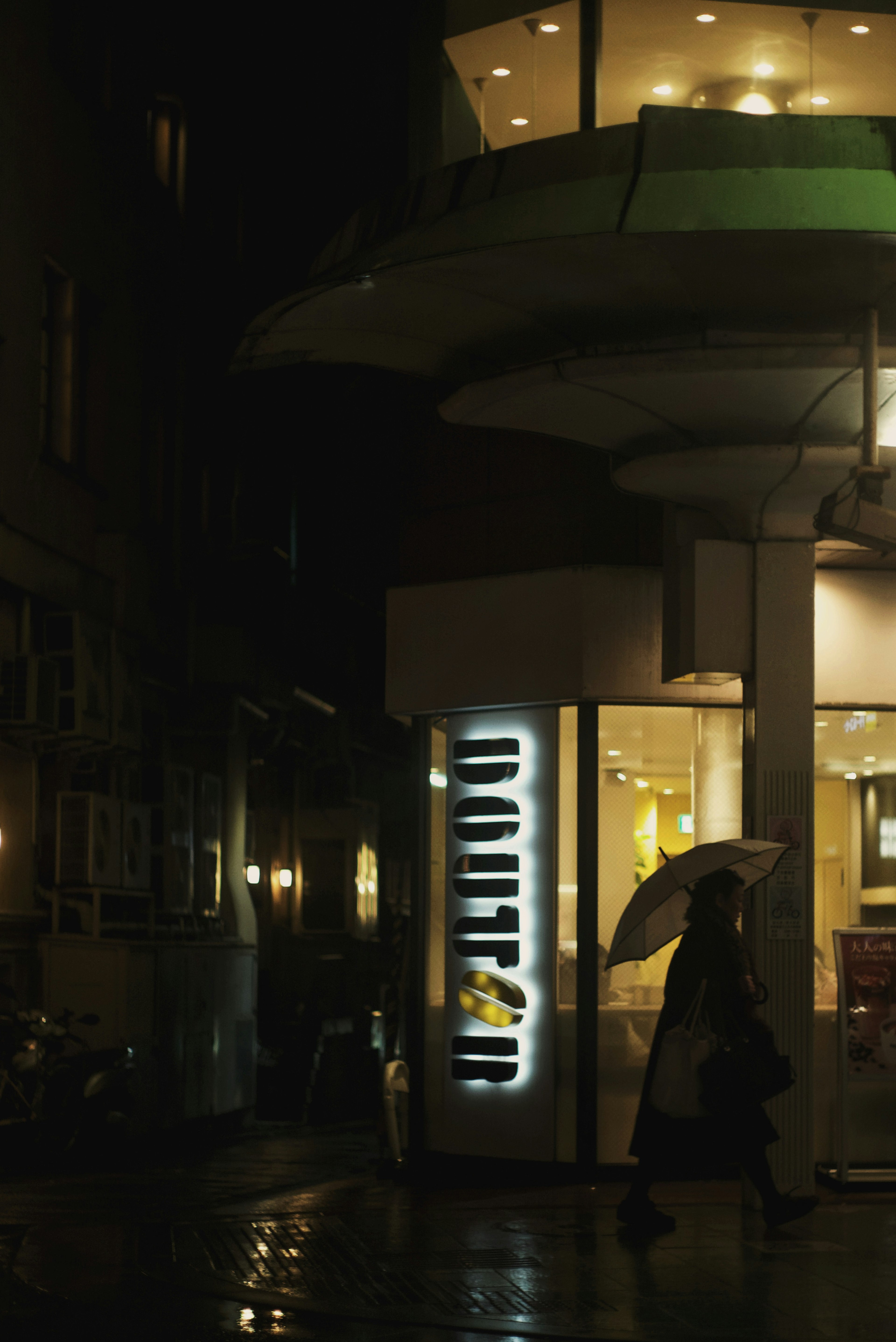 Person with an umbrella standing near a brightly lit sign in a dark city corner