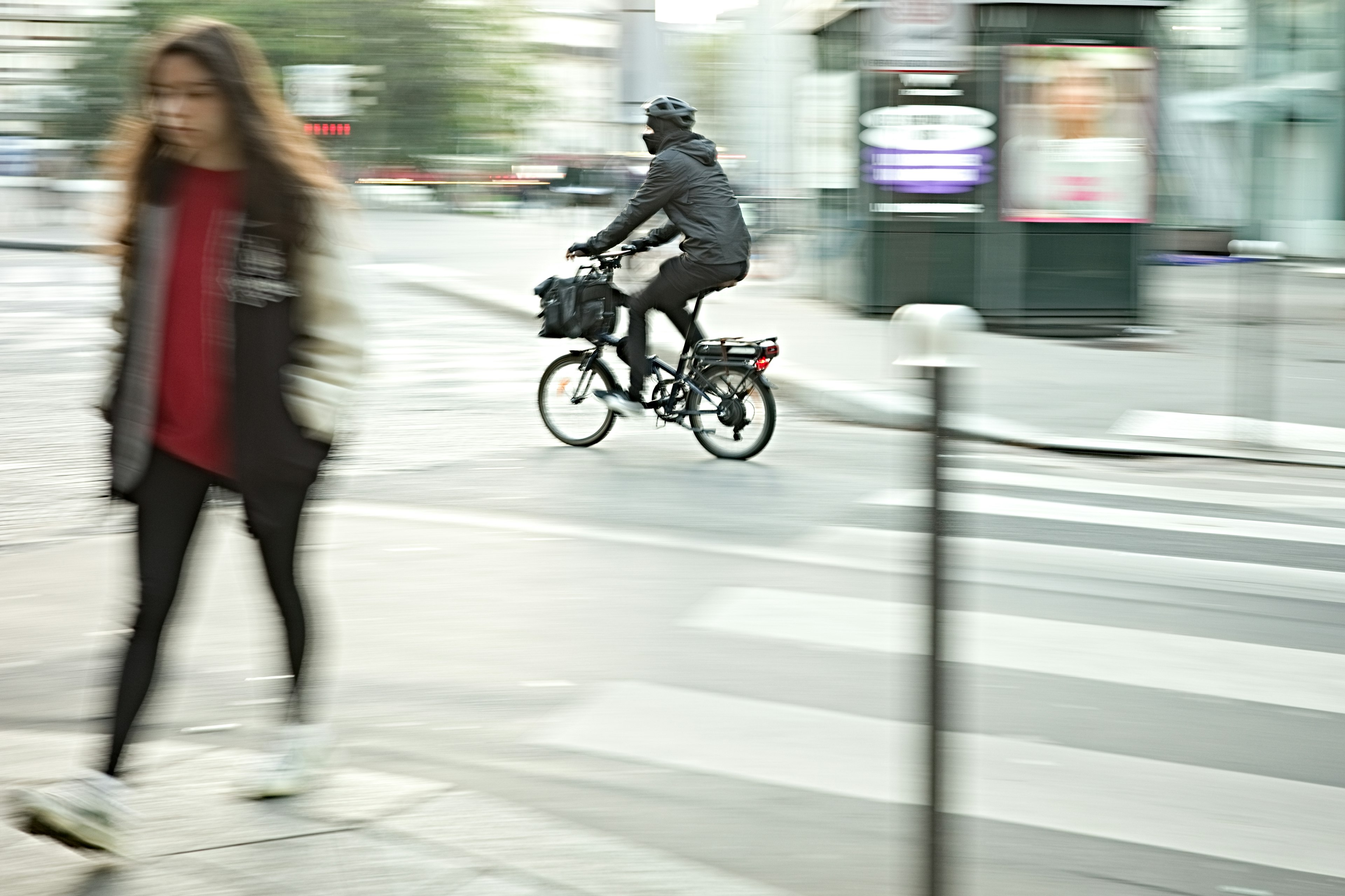 A delivery person on a bicycle crossing the street while a woman in a red shirt walks by