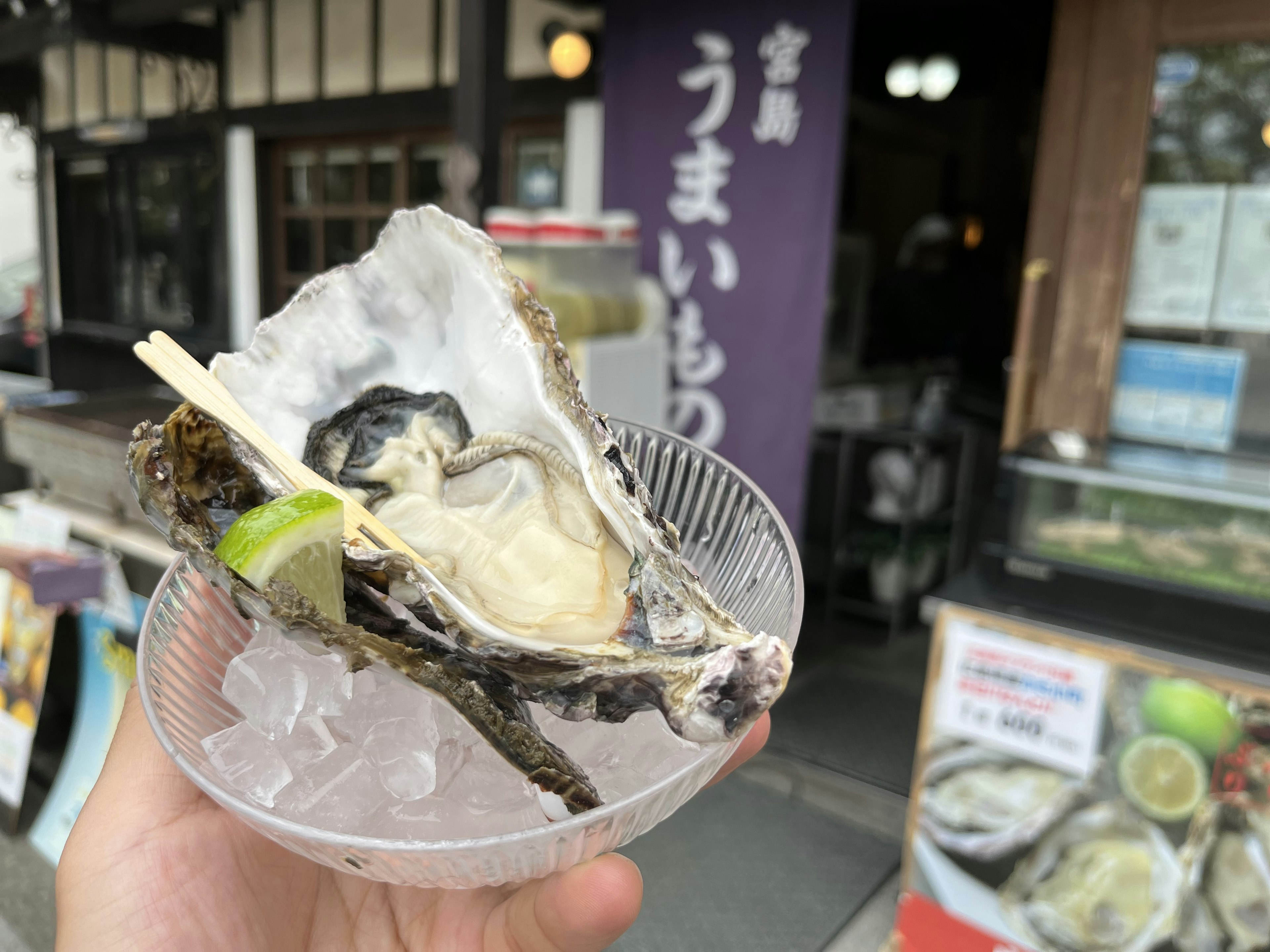 Une main tenant une huître fraîche avec une tranche de citron vert dans un plat transparent devant un panneau de restaurant japonais