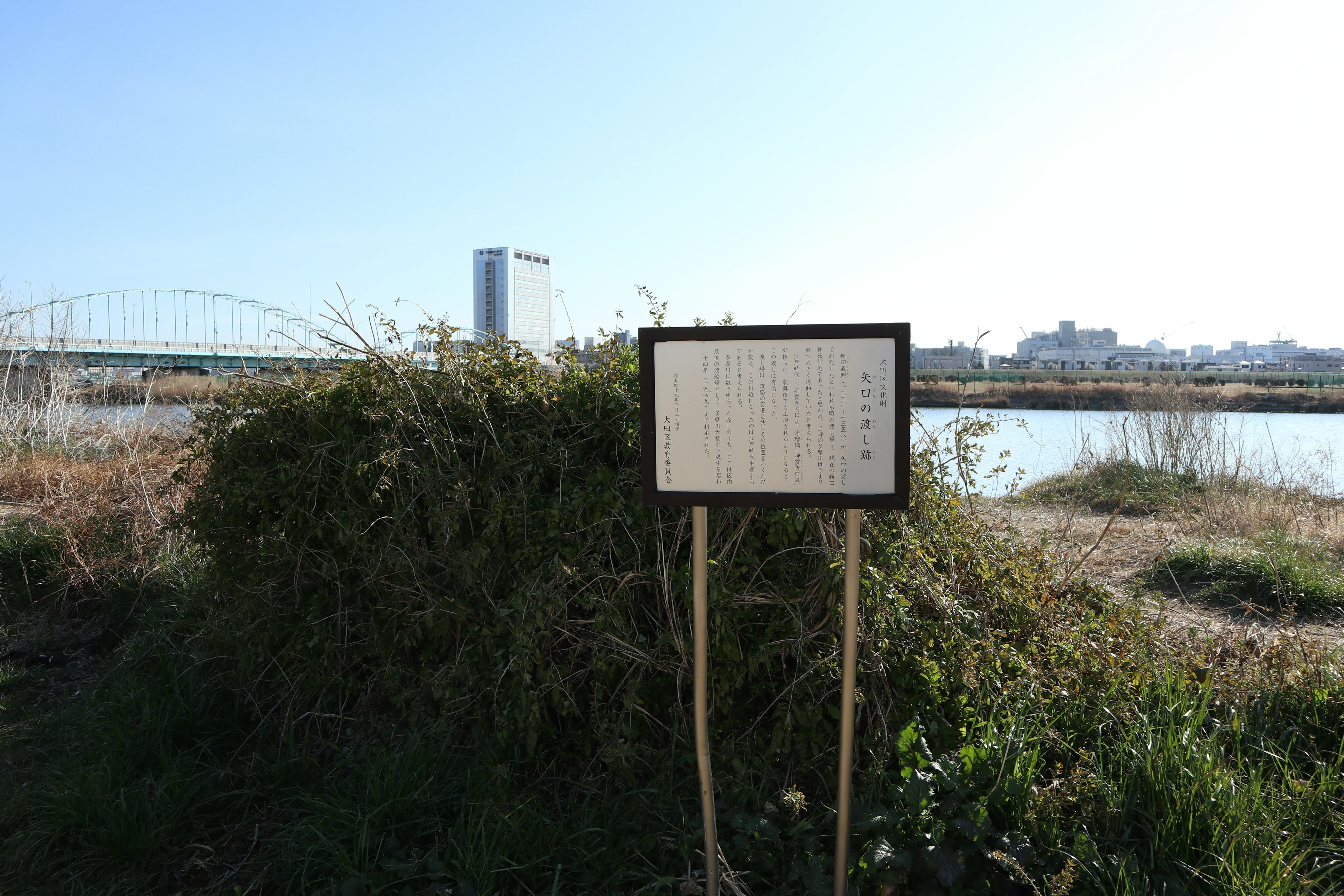 A sign standing in a grassy area by the river with a distant building