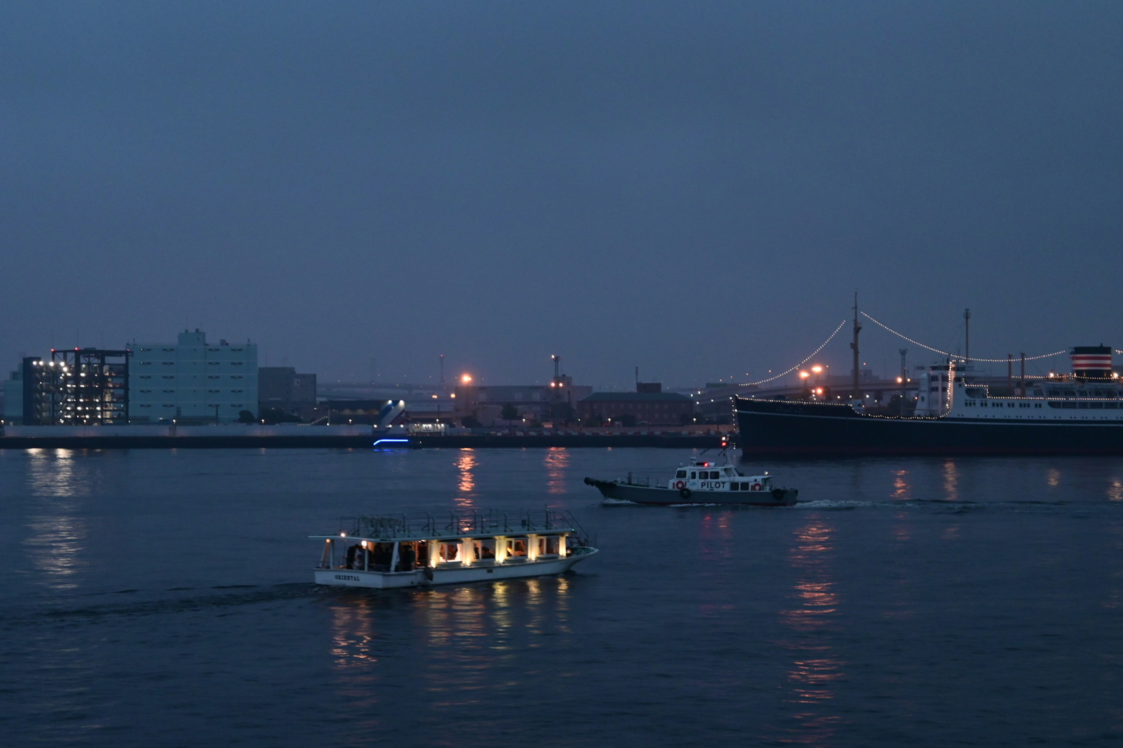 Vista nocturna de un barco navegando en el puerto con edificios iluminados y un gran barco