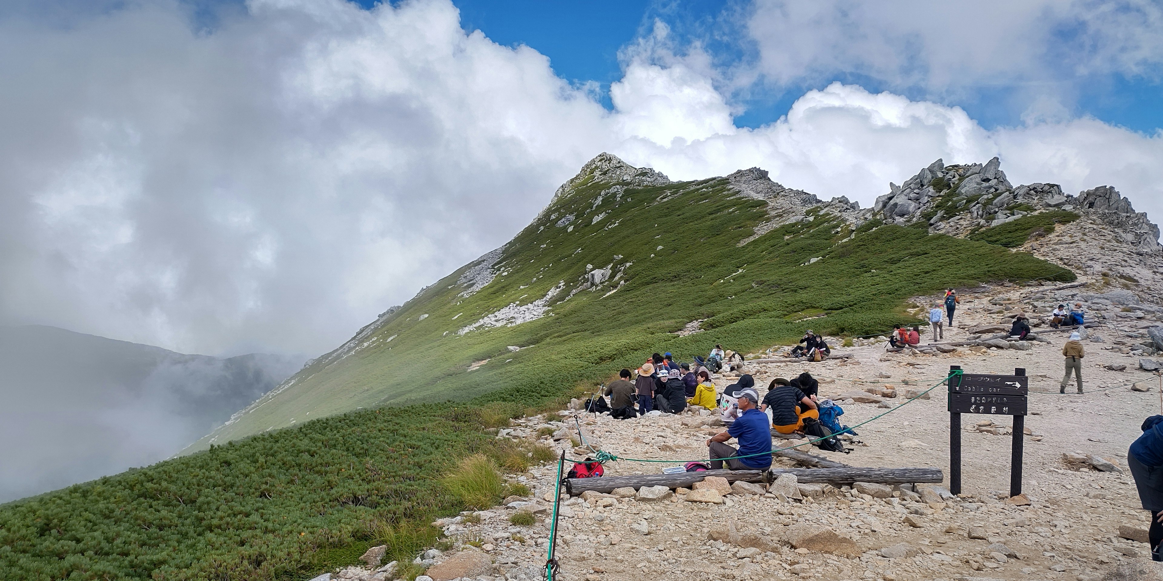 Foto de un paisaje montañoso con excursionistas reunidos en un área escénica con cielo azul y nubes