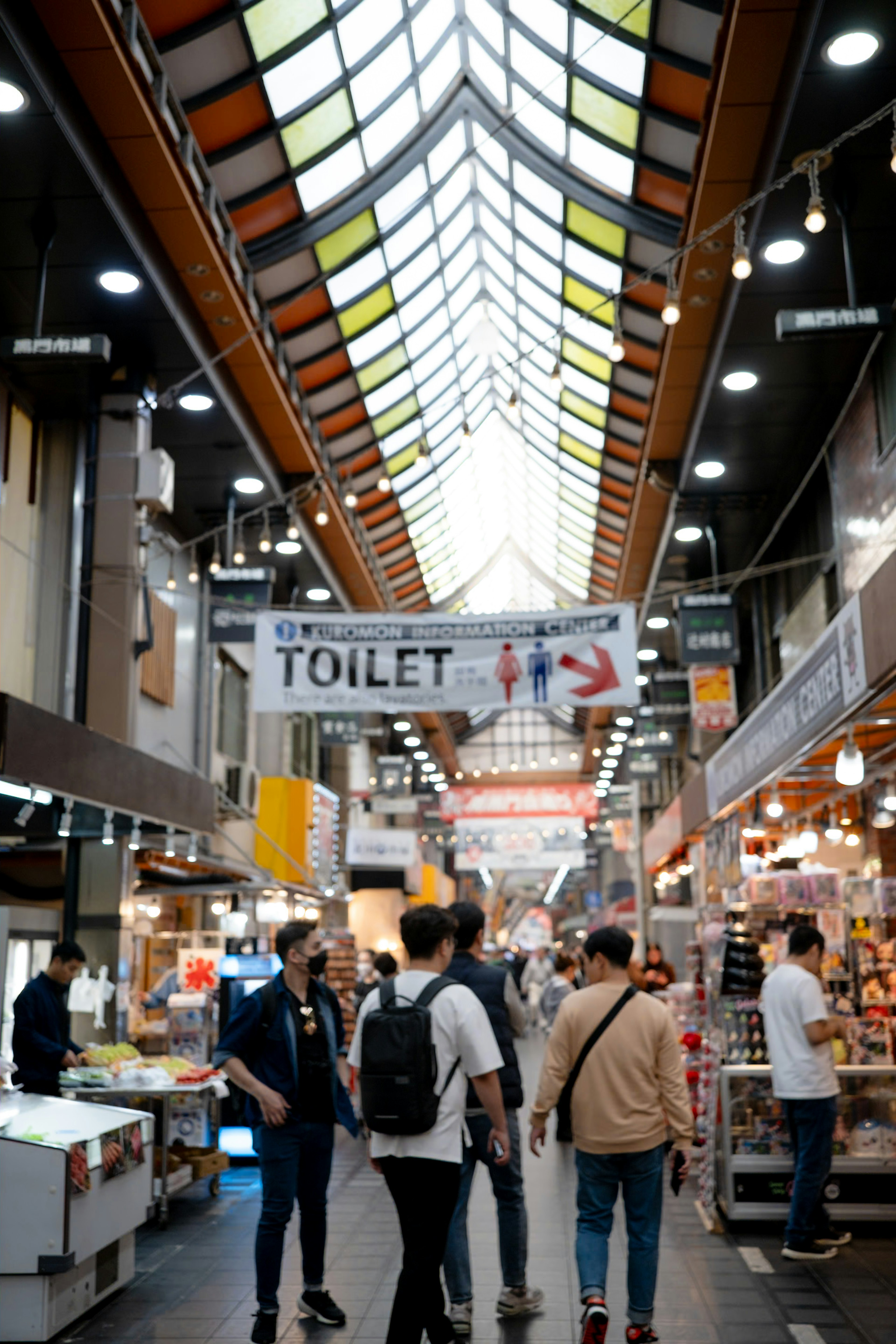 Un gruppo di persone all'interno di un mercato con illuminazione brillante e un design di tetto distintivo con un segnale di toilette ben visibile