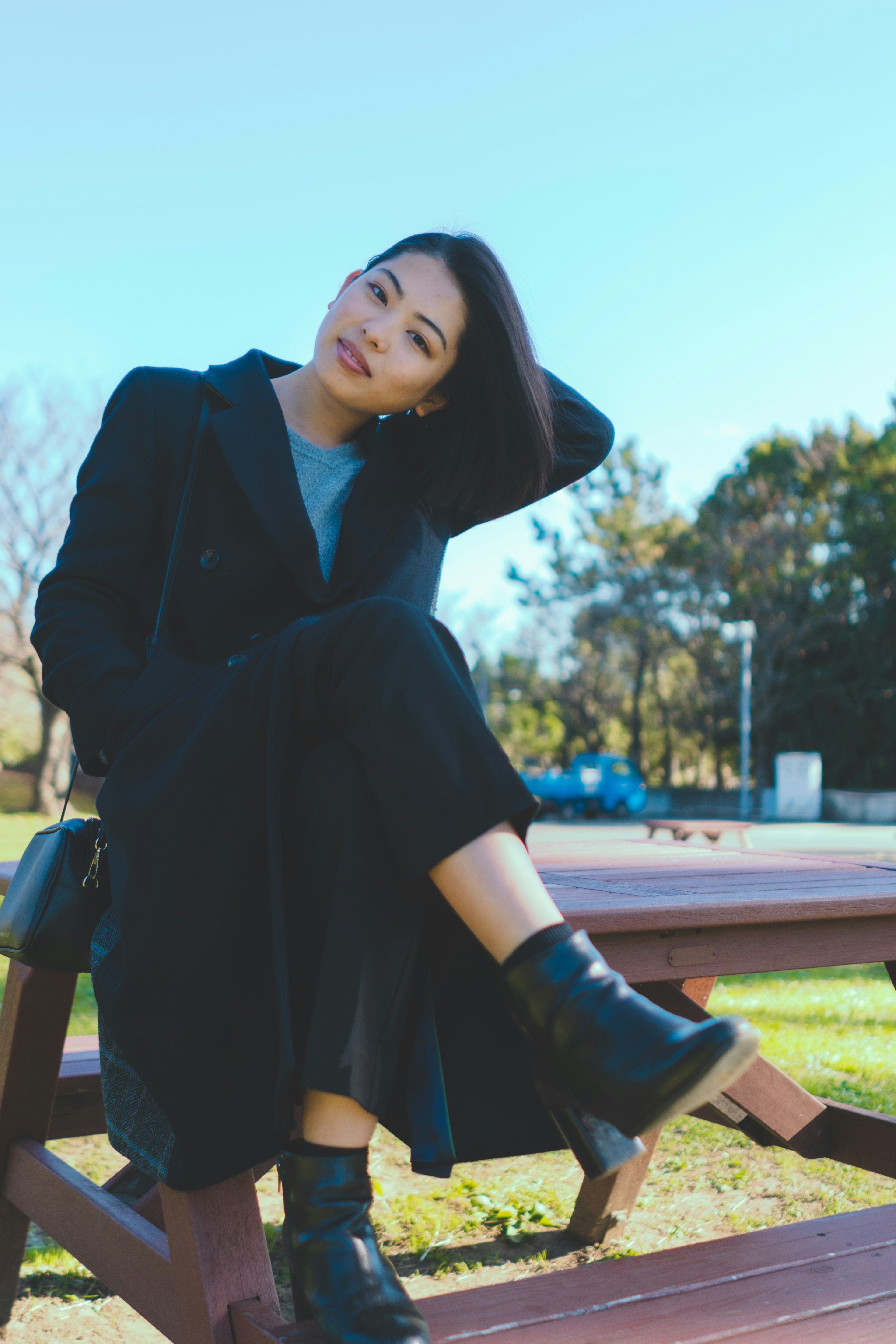 Portrait of a woman sitting on a park bench wearing a black coat and boots smiling while adjusting her hair