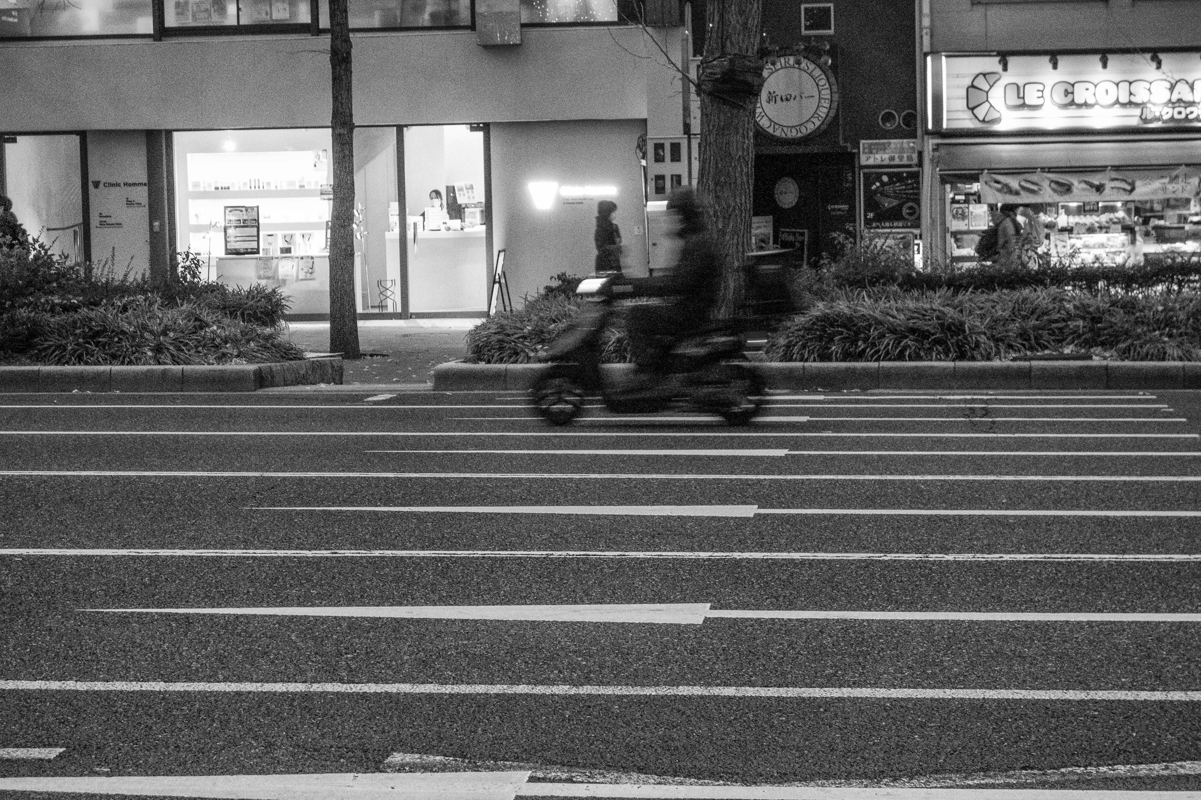 Une moto passant par un passage piéton dans une scène urbaine en noir et blanc avec des magasins