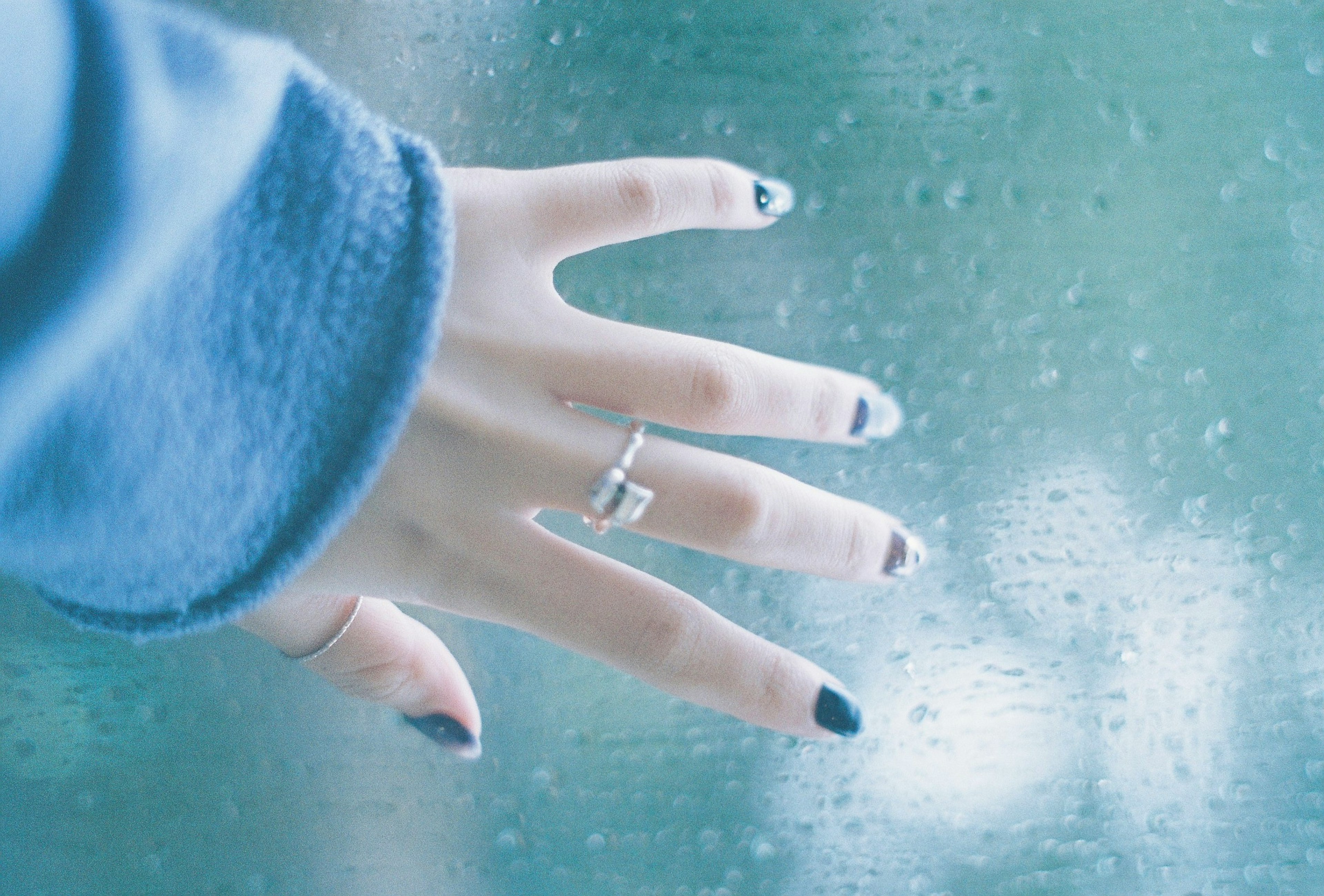 Primo piano di una mano che tocca una finestra bagnata dalla pioggia con un maglione blu