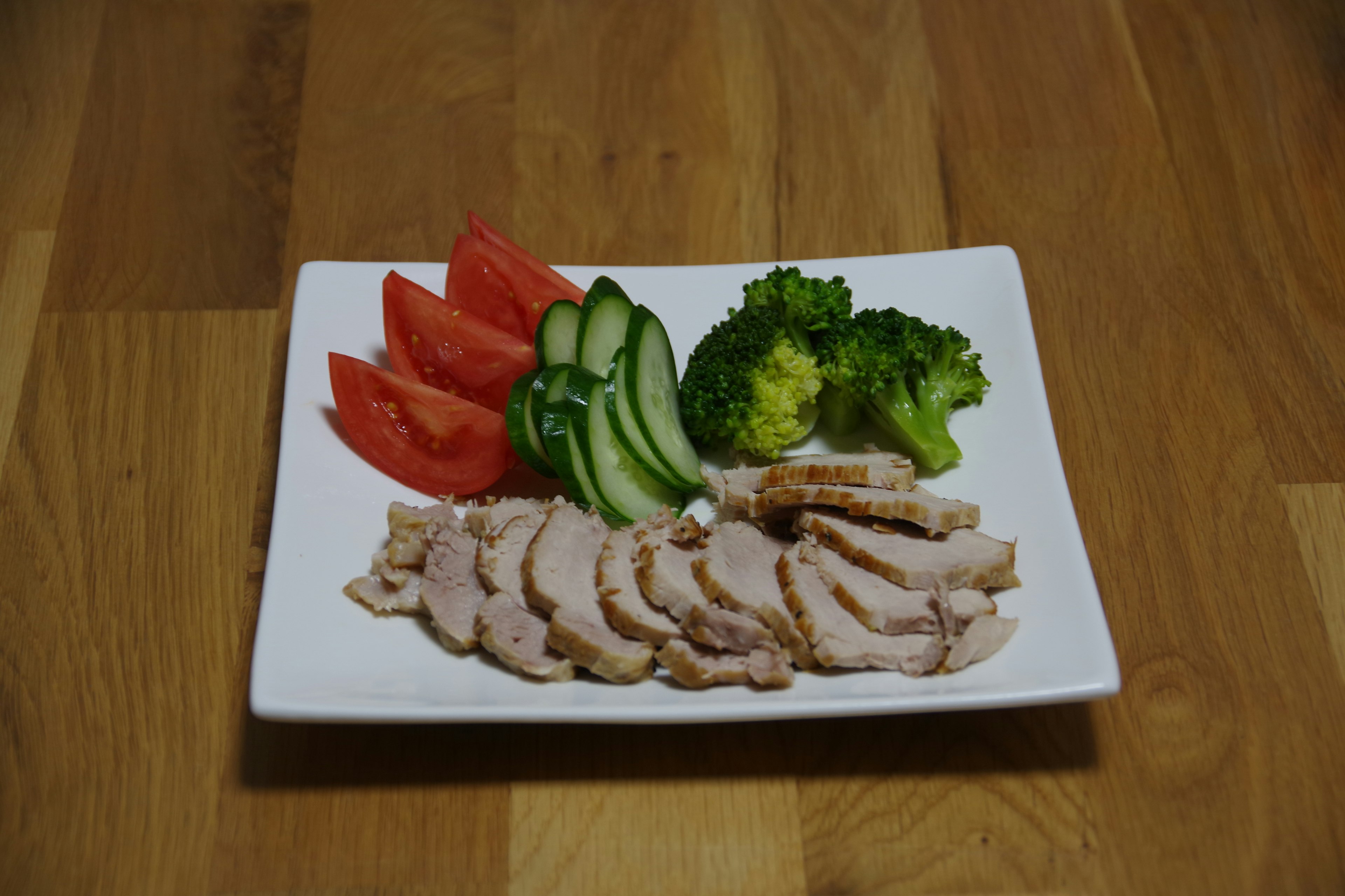 Sliced chicken with tomatoes, cucumbers, and broccoli on a white plate