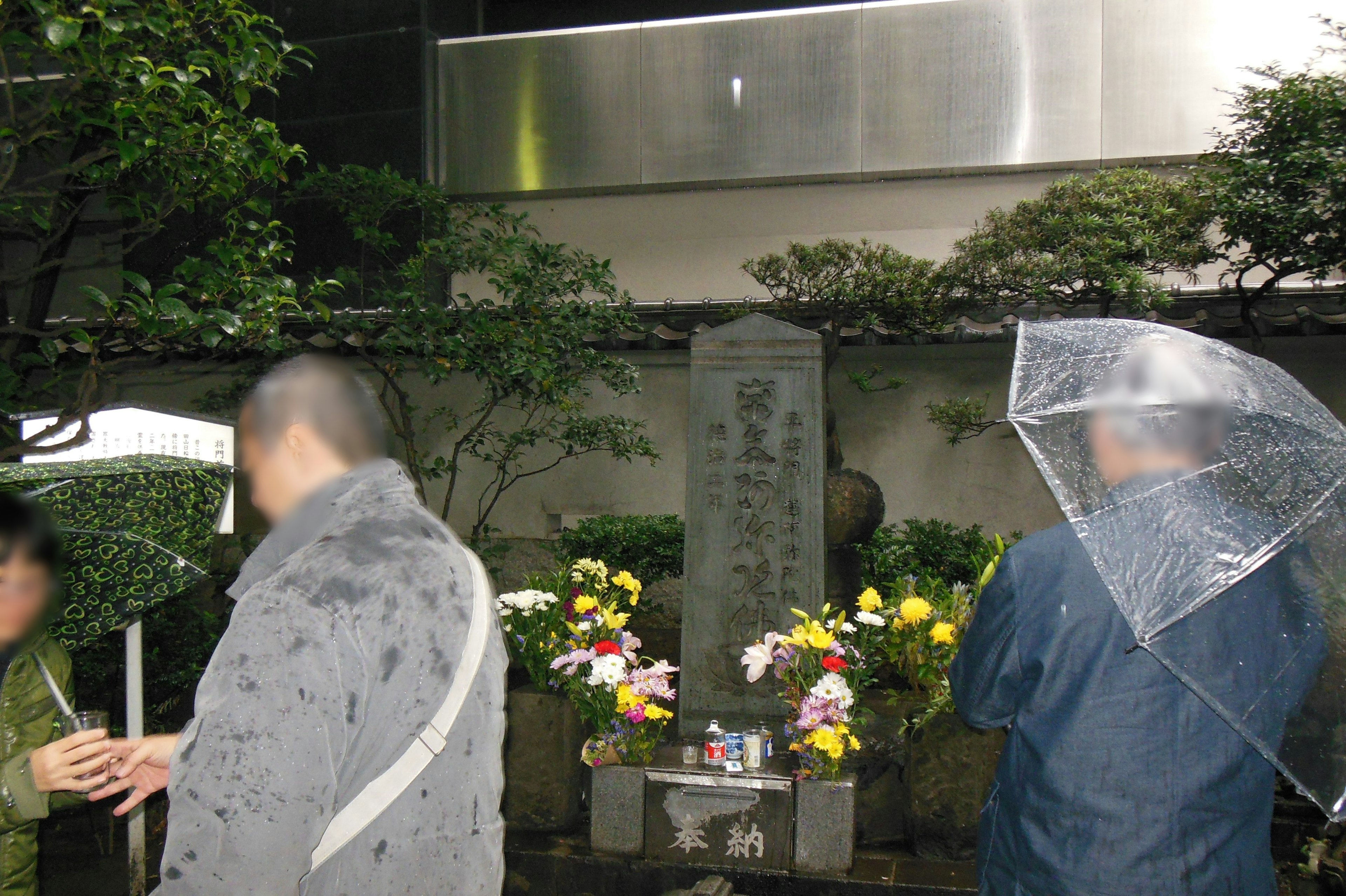 People standing in the rain near a stone monument adorned with flowers