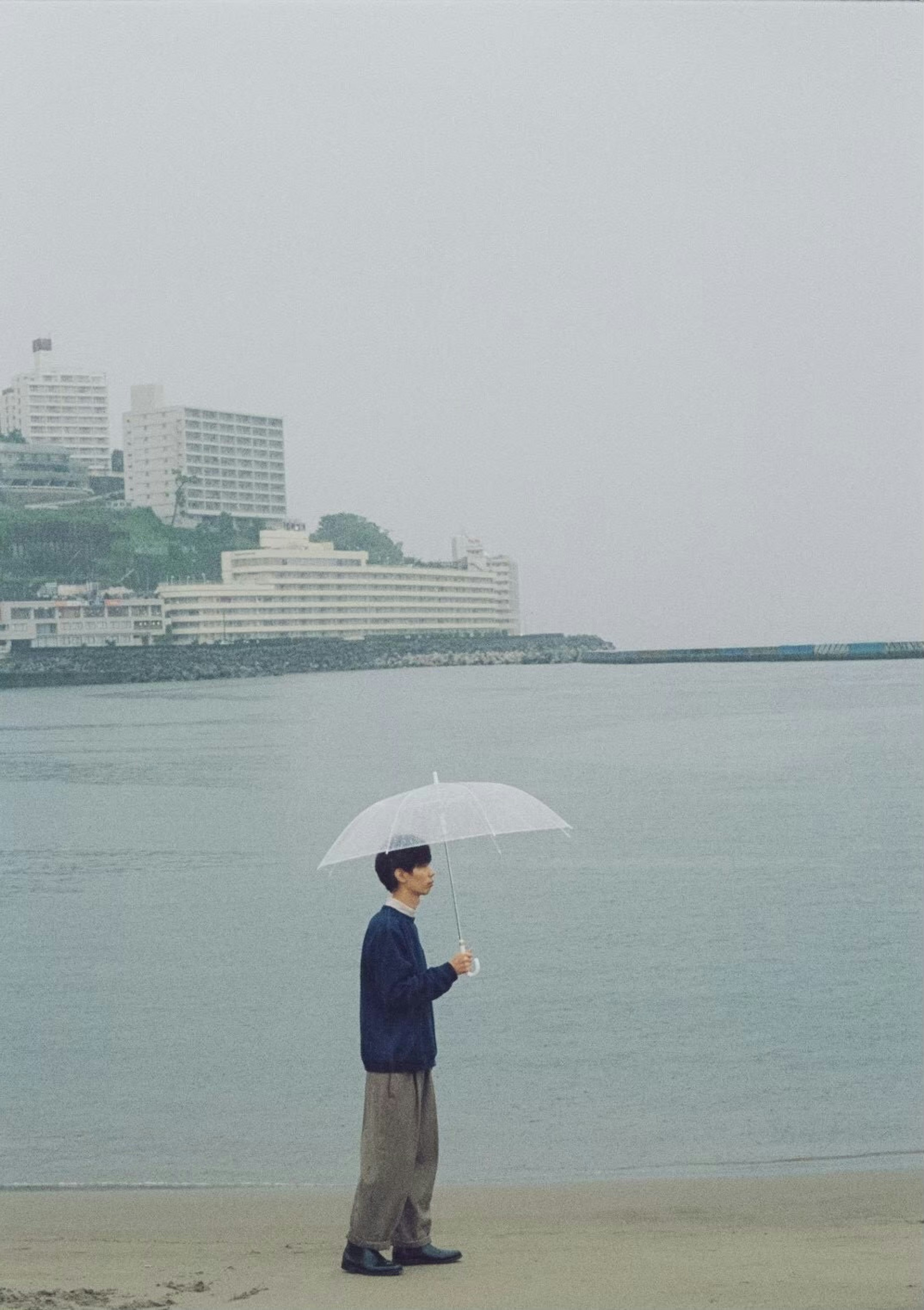 Mann mit Regenschirm am Meer mit Gebäuden im Hintergrund und Nebel über dem Wasser