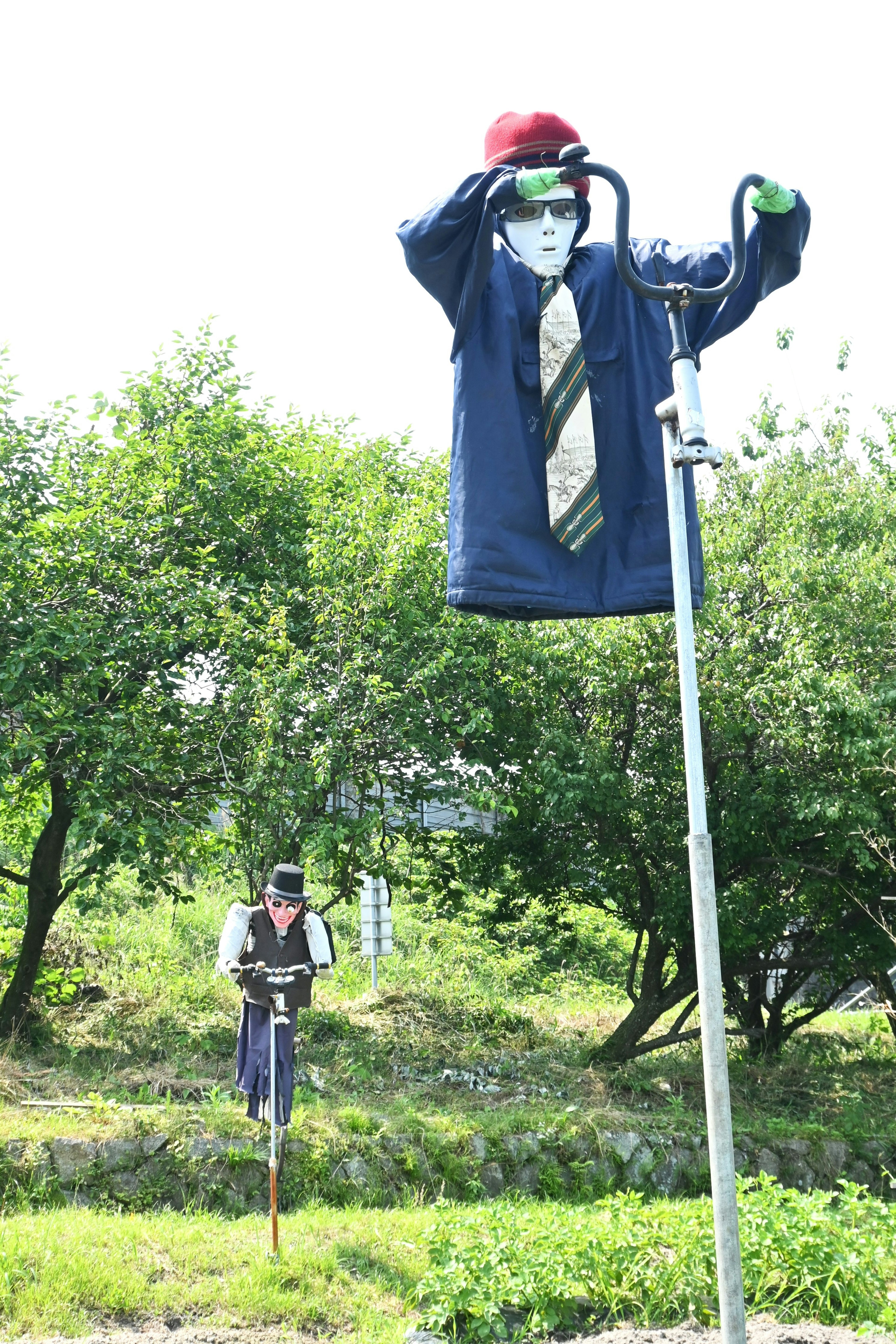 A scarecrow-like figure on a tall pole wearing a blue suit and red hat with raised hands behind another figure standing still