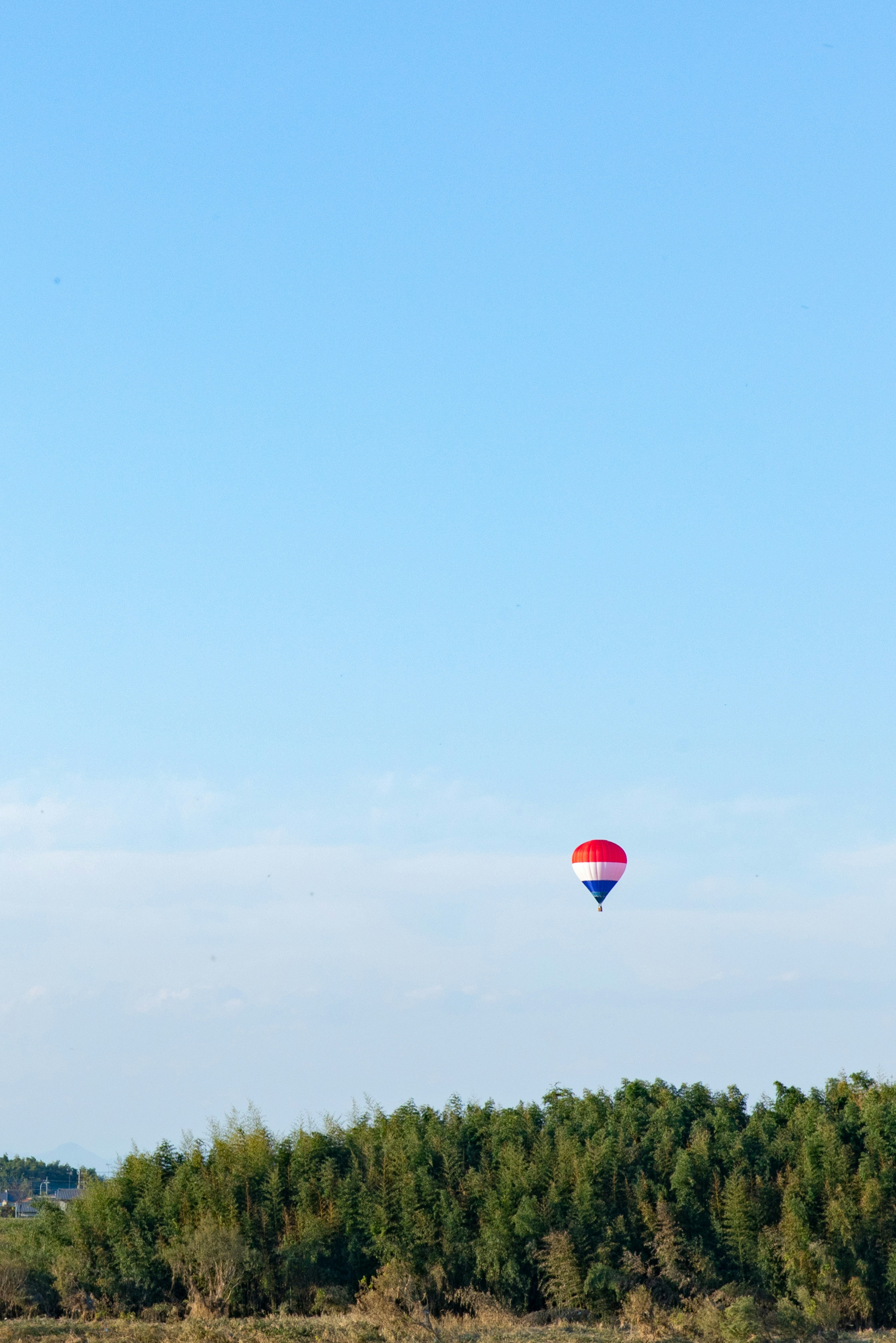 Red and white hot air balloon floating in a blue sky above green trees