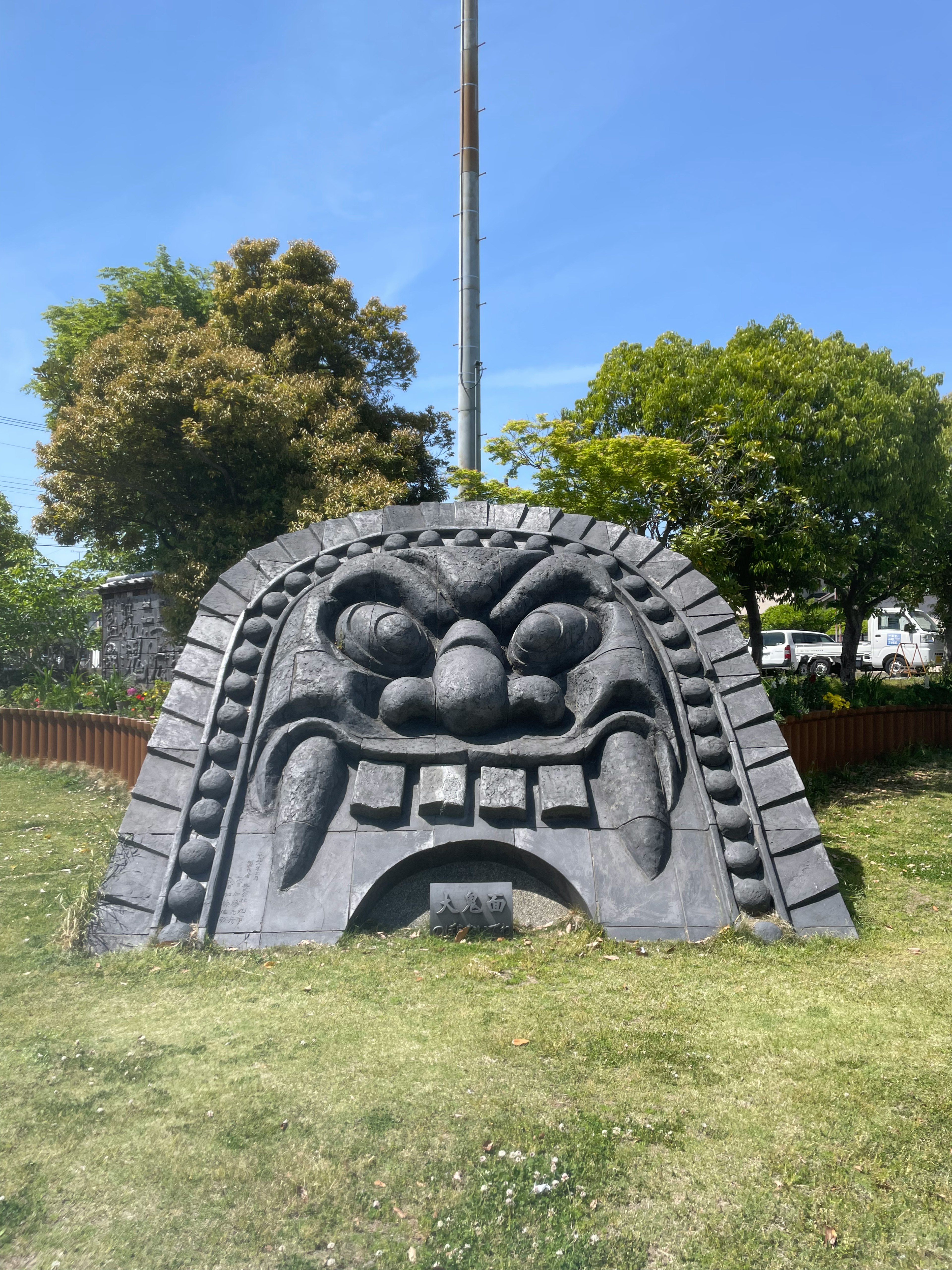 公園にある大きな石の彫刻の顔　目が大きくて口が開いている　周りに緑の木々と青い空が見える