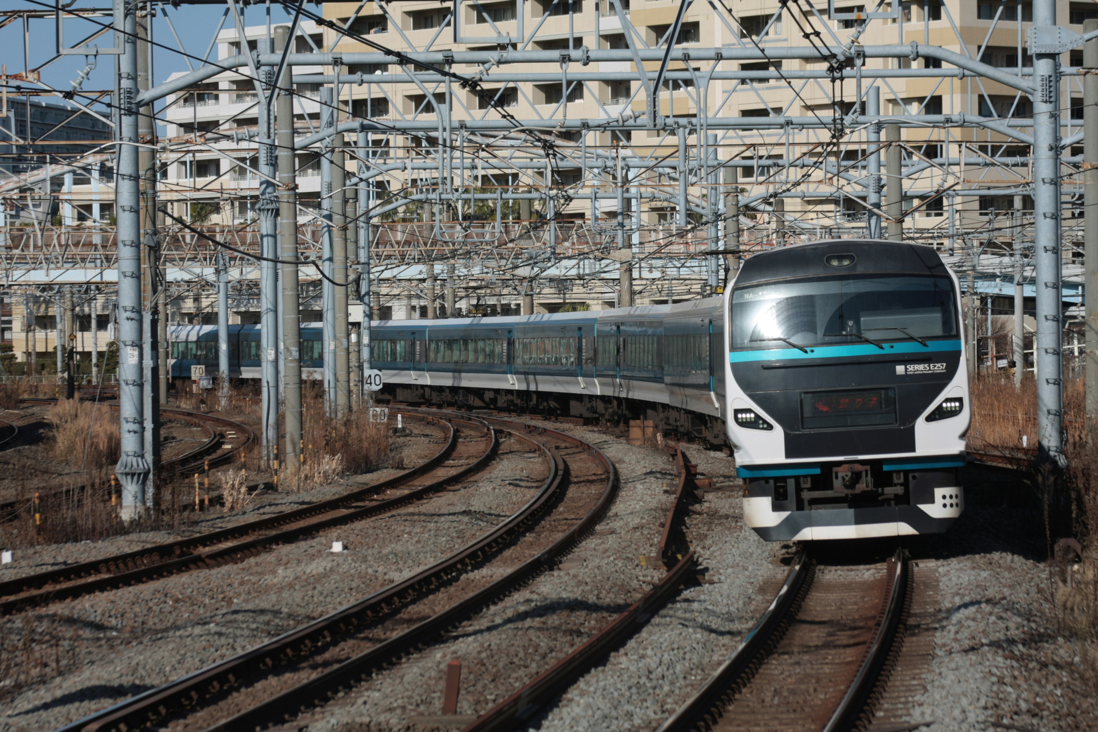 A train navigating curved tracks with overhead wires and nearby buildings in the background