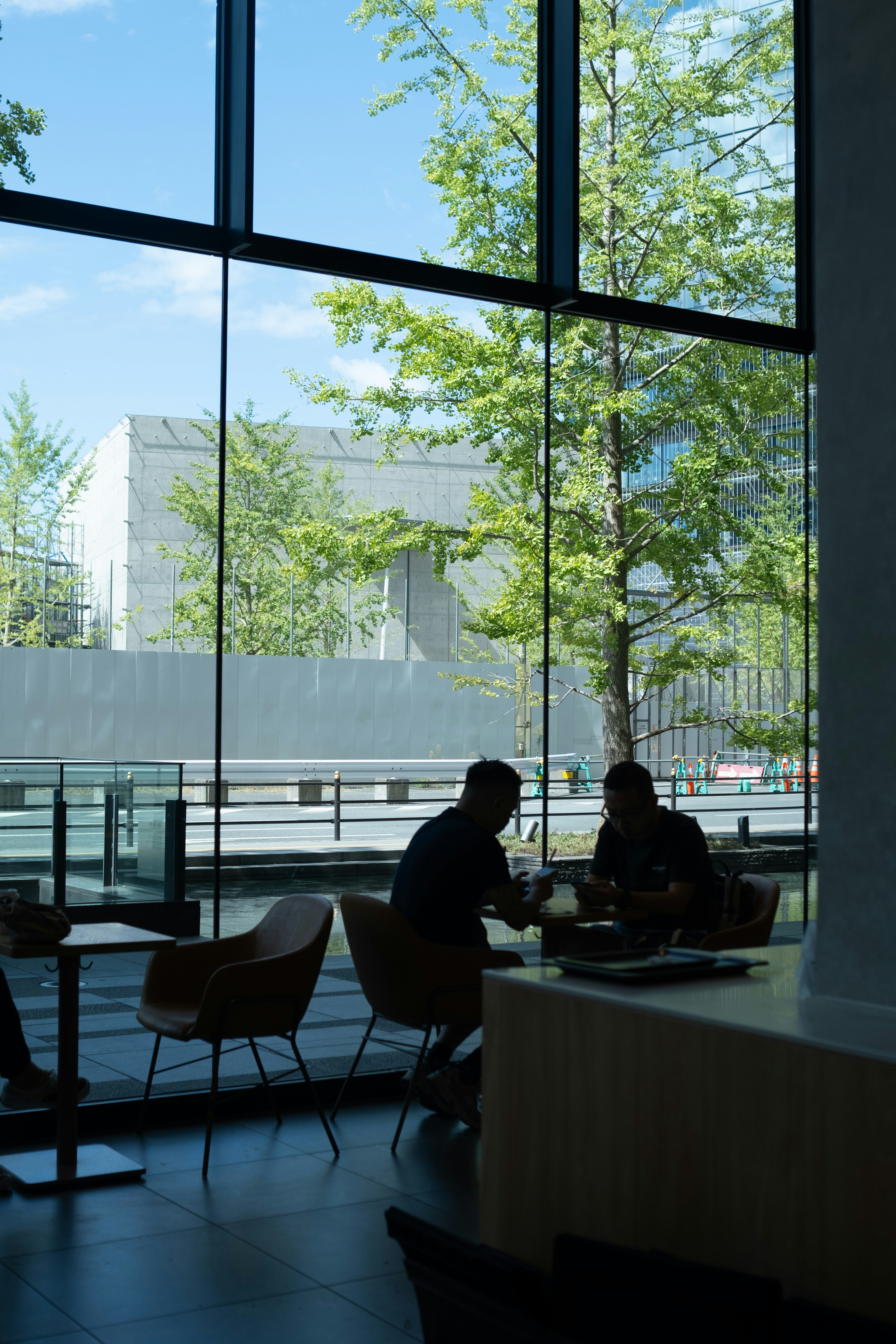 Zwei Personen sitzen in einem hellen Café und schauen durch große Fenster auf grüne Bäume draußen