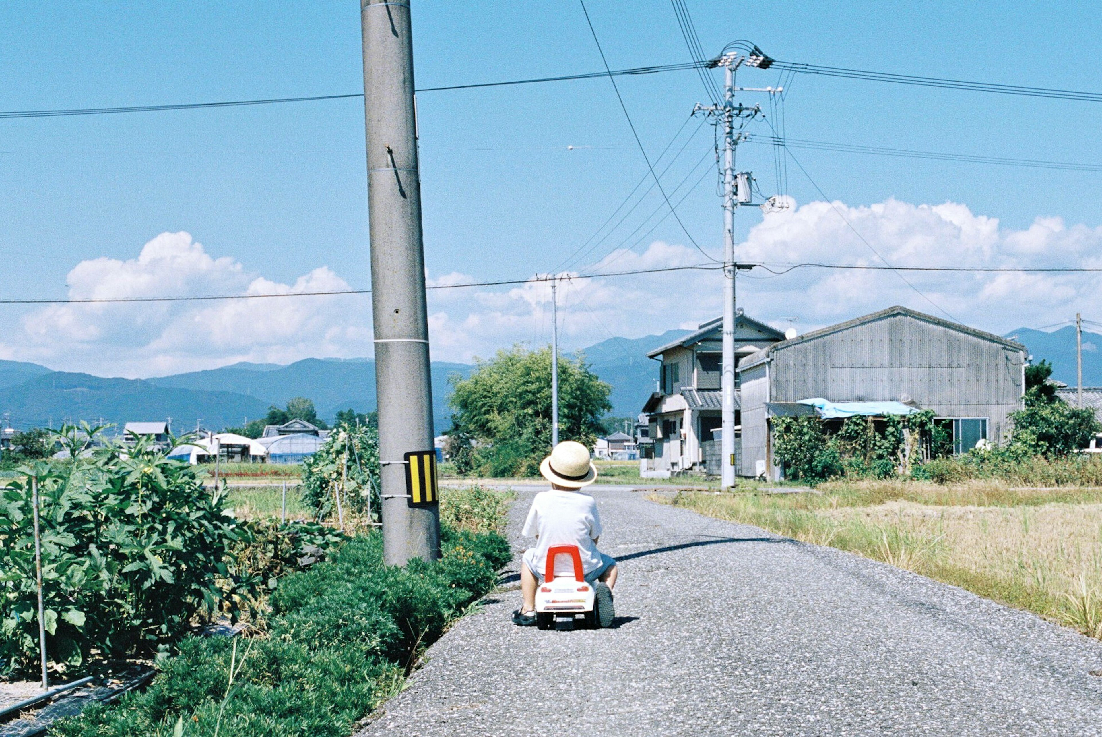 A child riding a small red toy car on a rural path under a blue sky