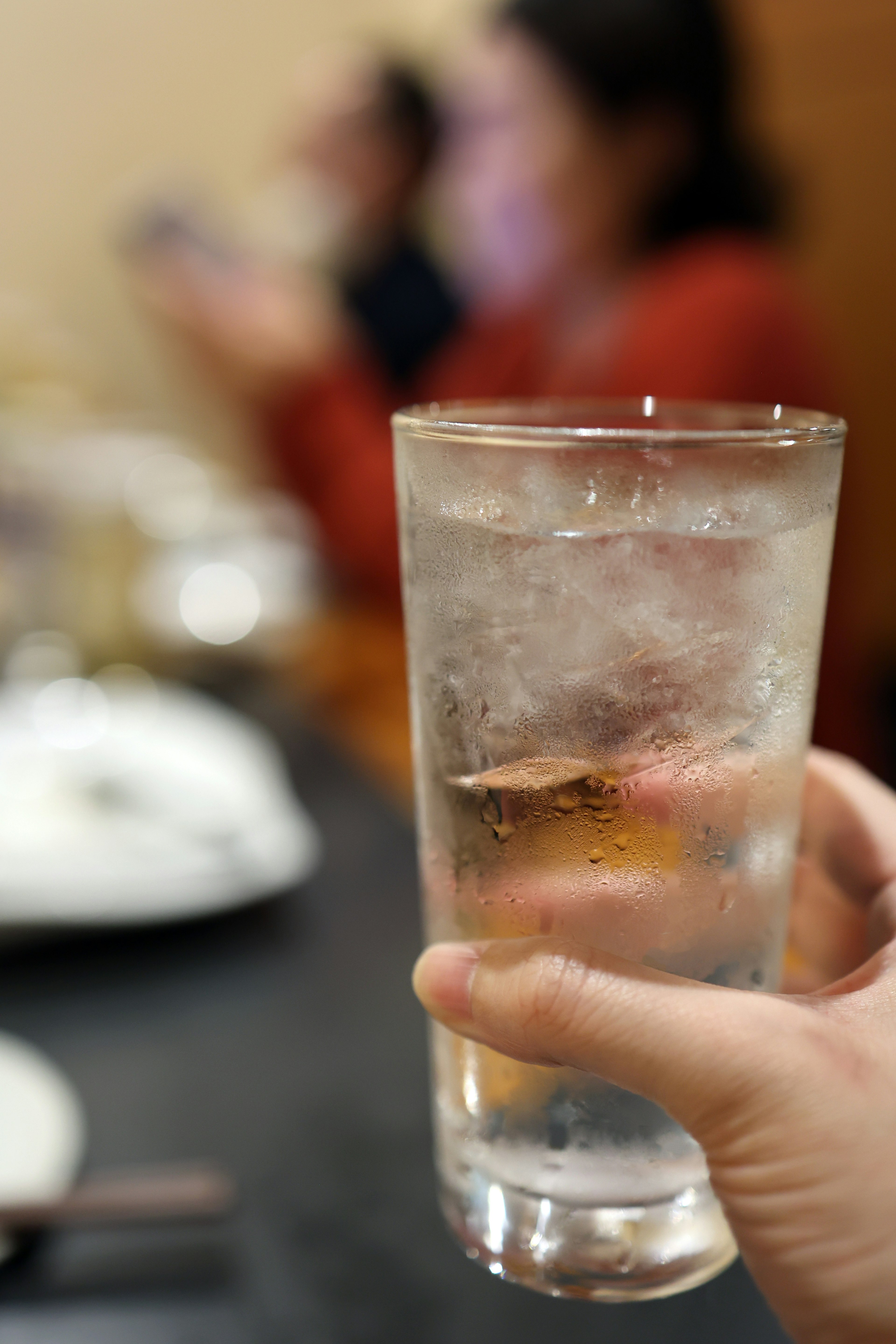 A hand holding a glass of ice water with blurred figures in the background