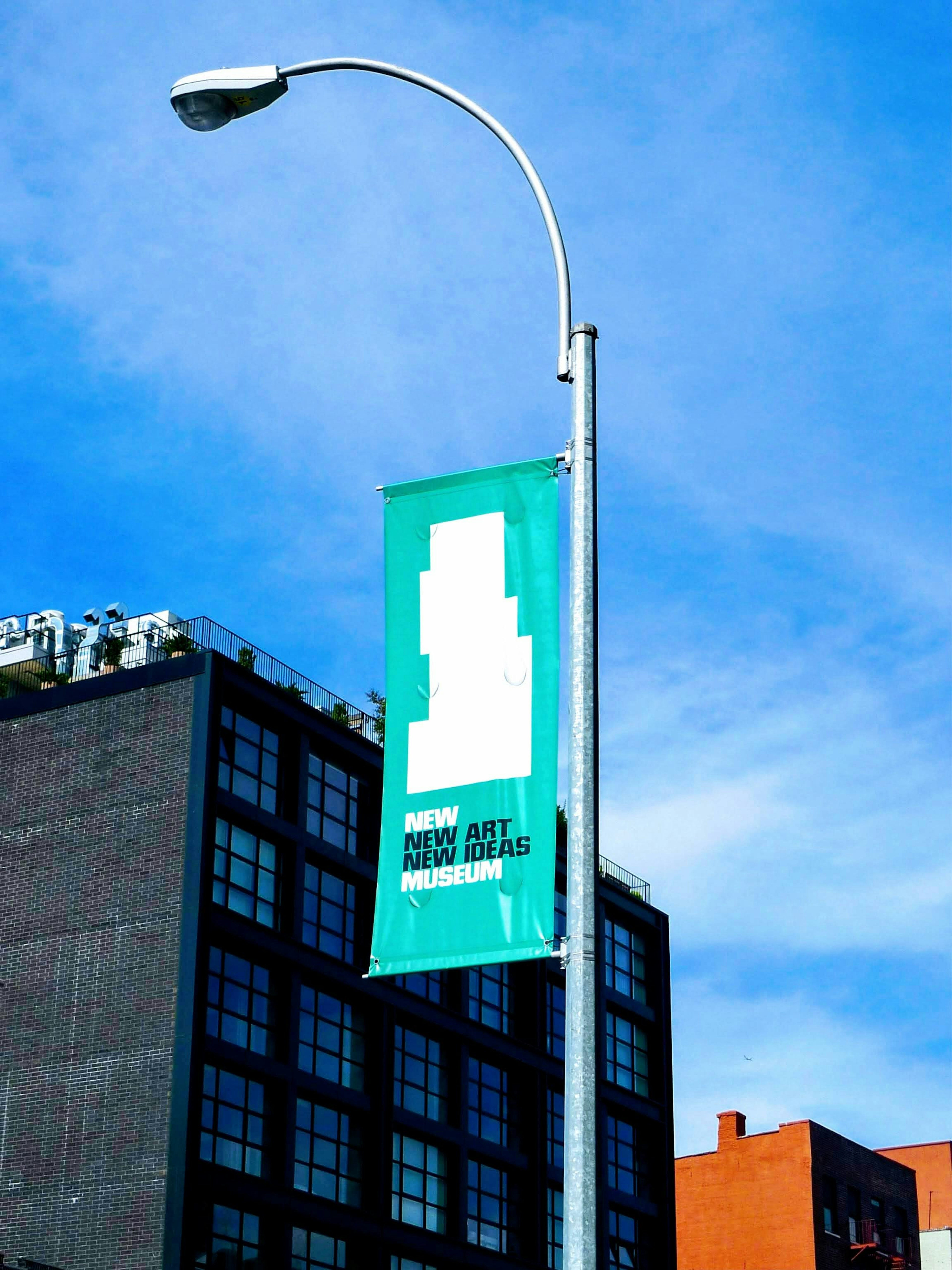 Green banner attached to a streetlight promoting a new art space under a blue sky