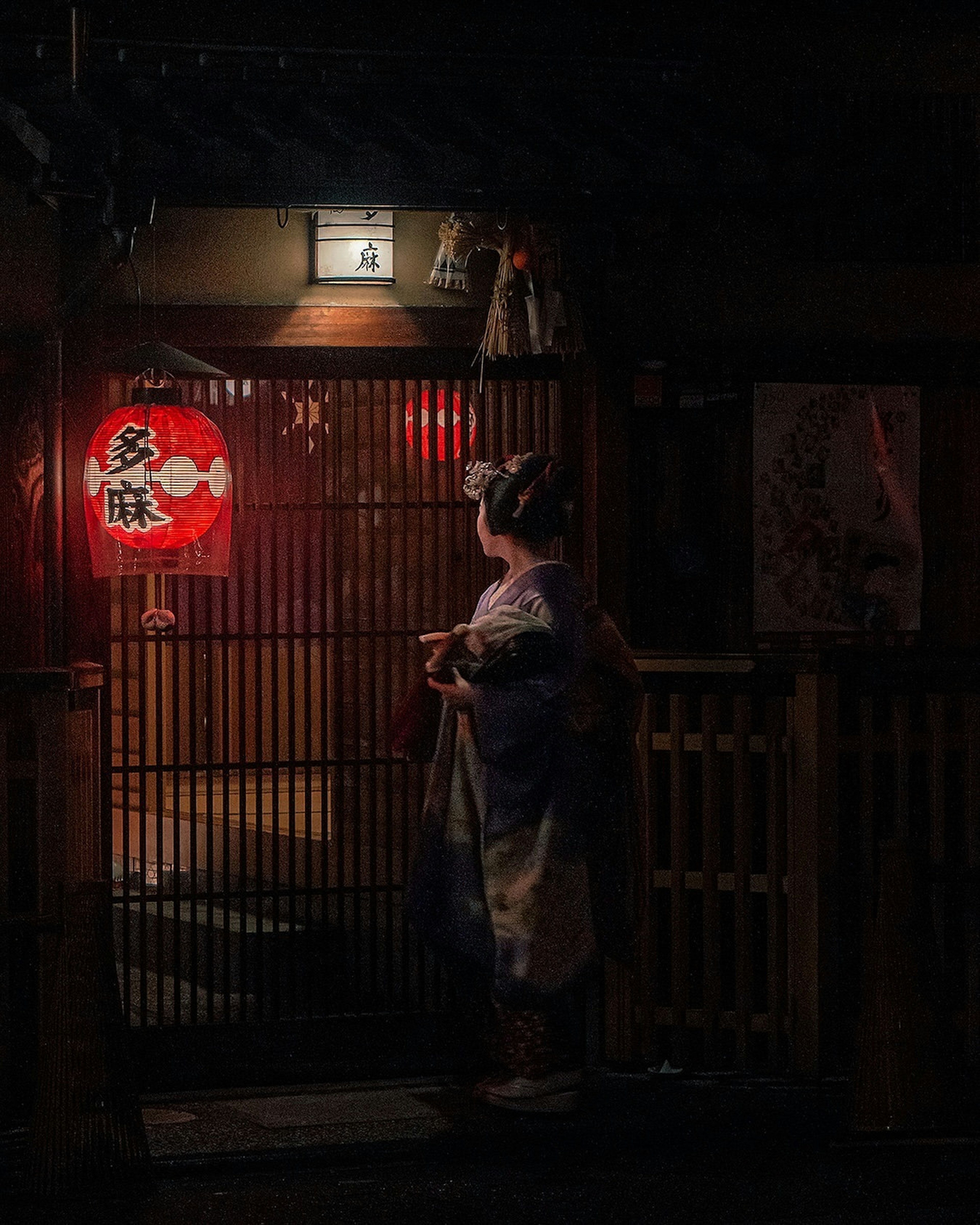 Una mujer en kimono de pie frente a una entrada japonesa tradicional con una linterna roja