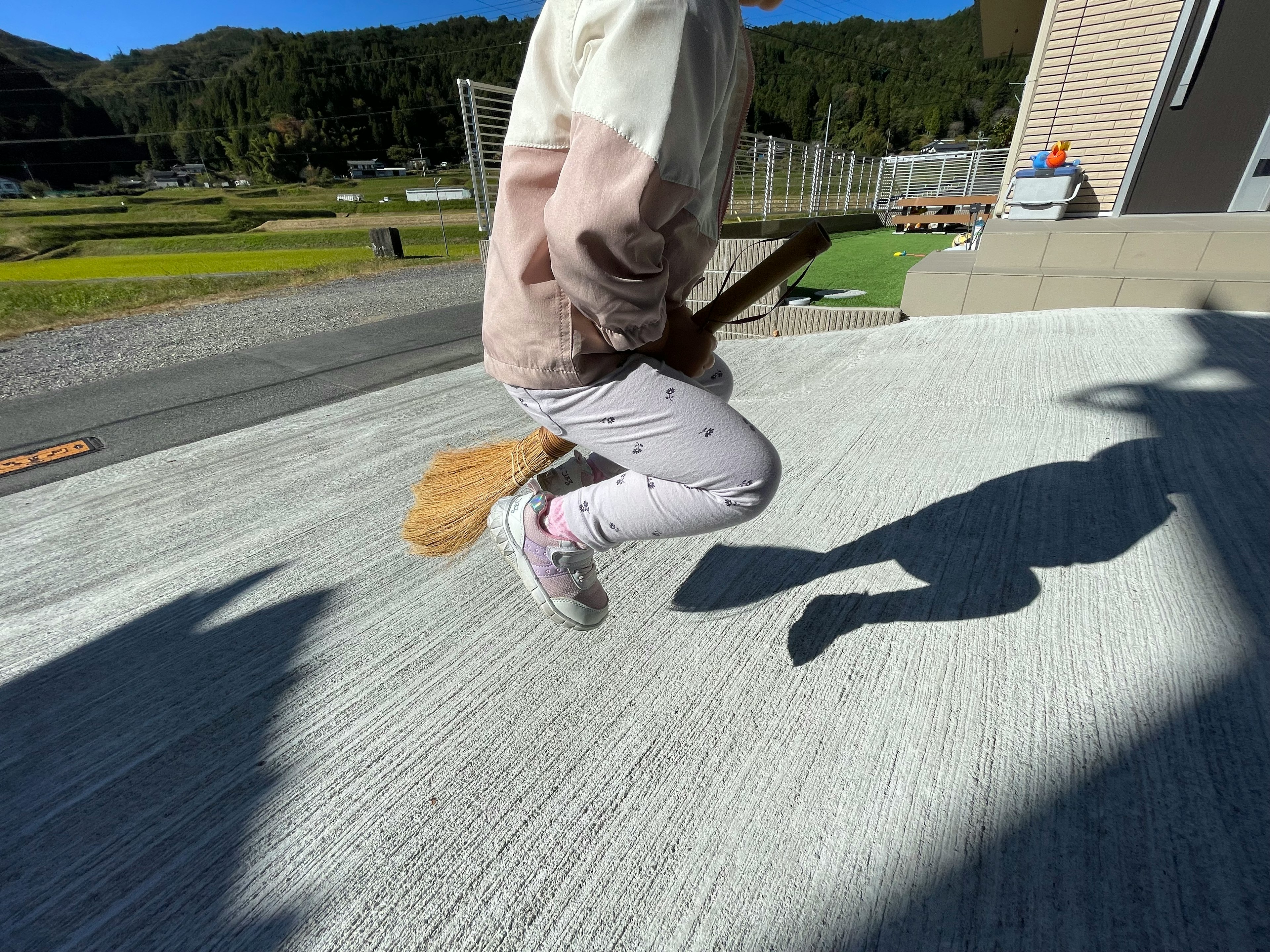 Child jumping with a broomstick-like object in a natural setting with a house in the background