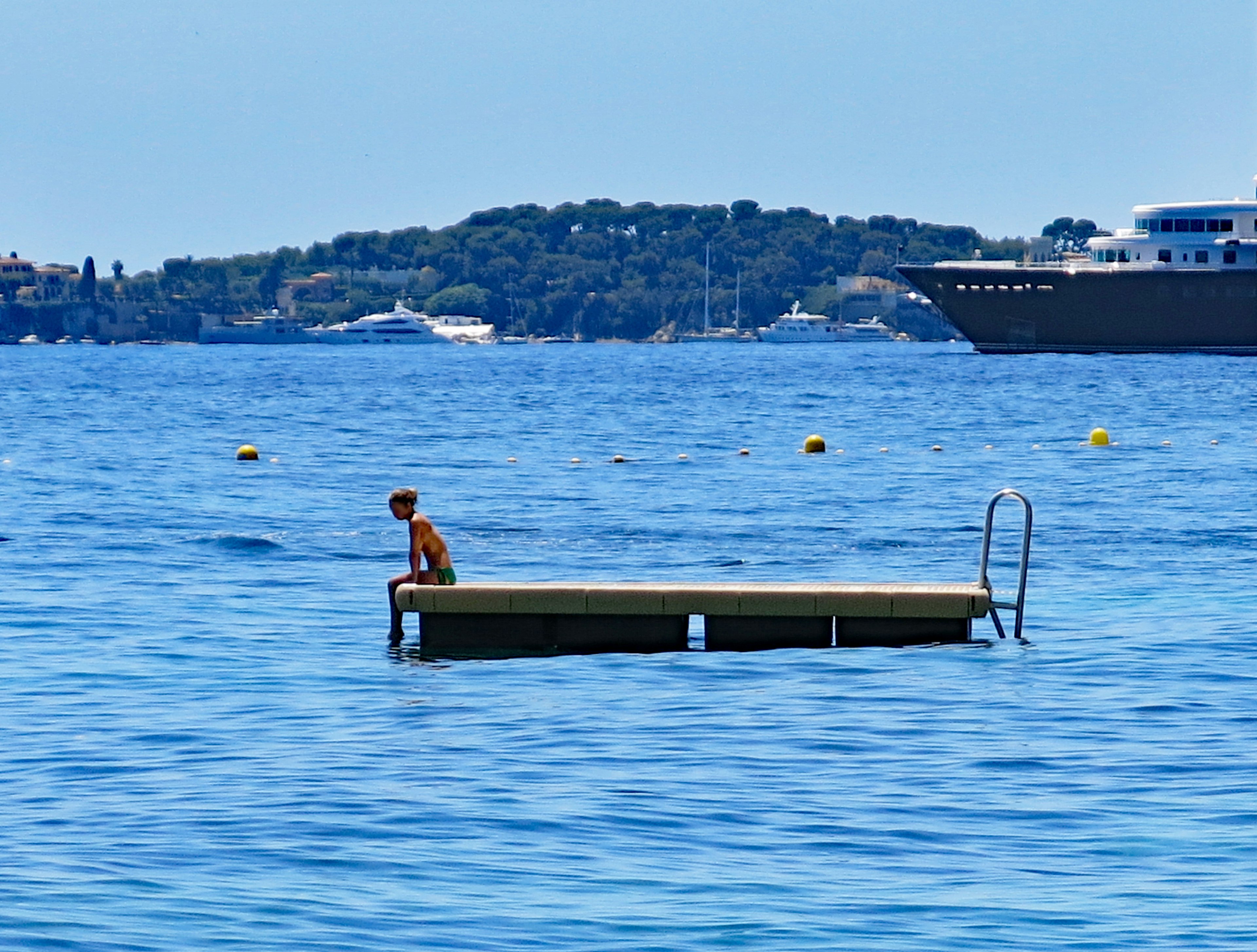 Ein Junge sitzt auf einem schwimmenden Steg im blauen Wasser mit einem Luxus-Yacht im Hintergrund