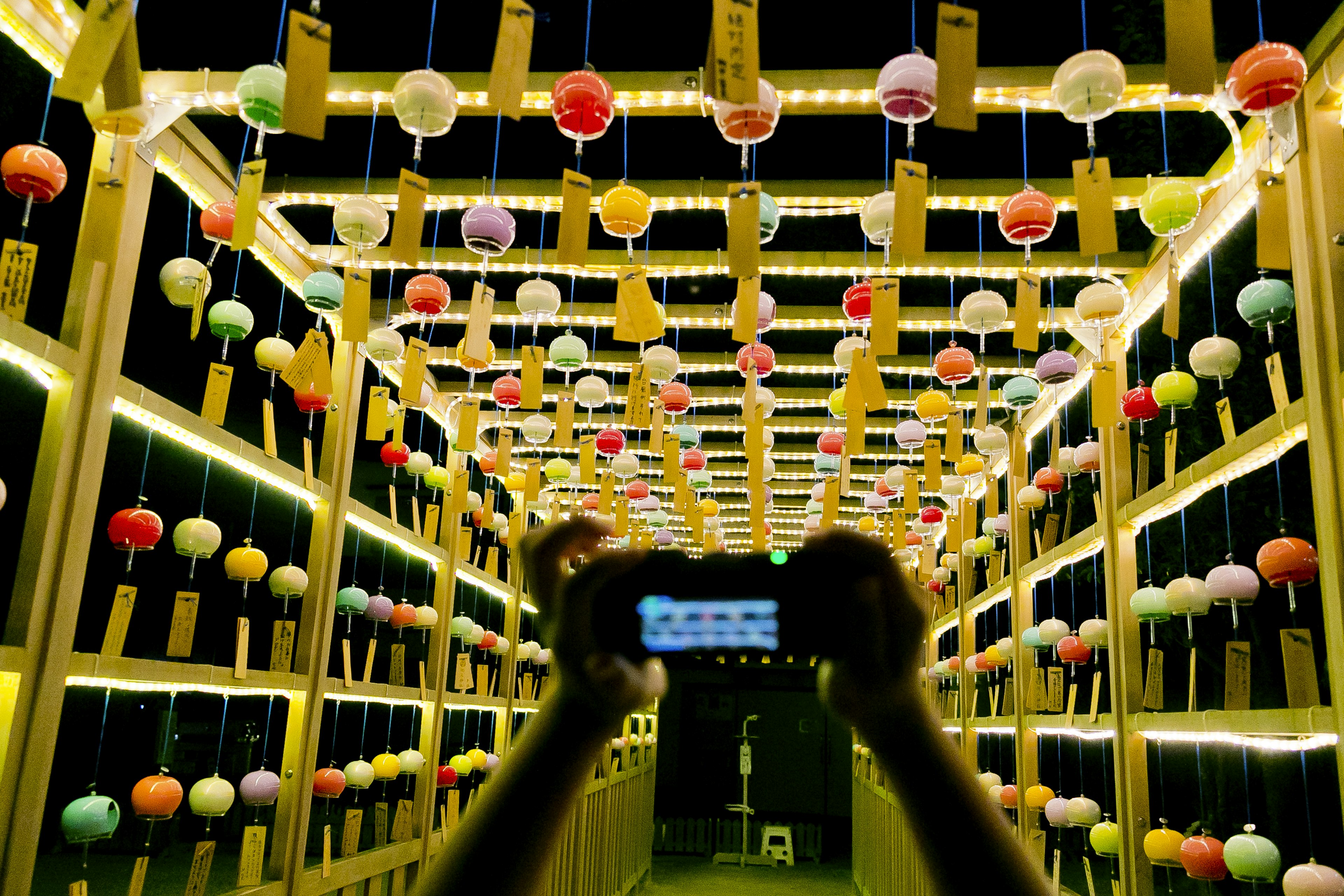 Hands holding a smartphone in a brightly lit tunnel adorned with colorful wind chimes