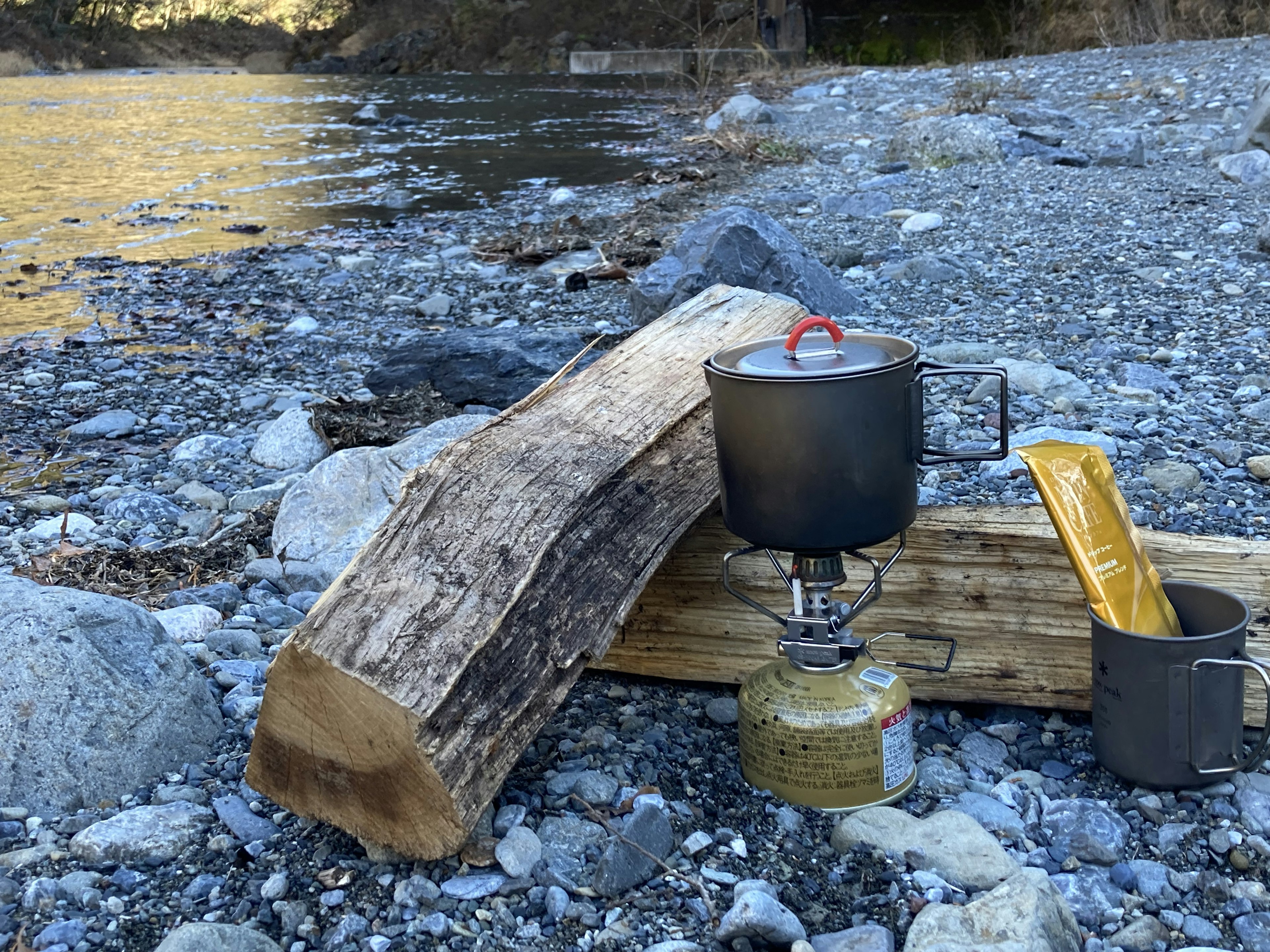 Estufa de camping y olla junto al río con un tronco grande y guijarros cerca