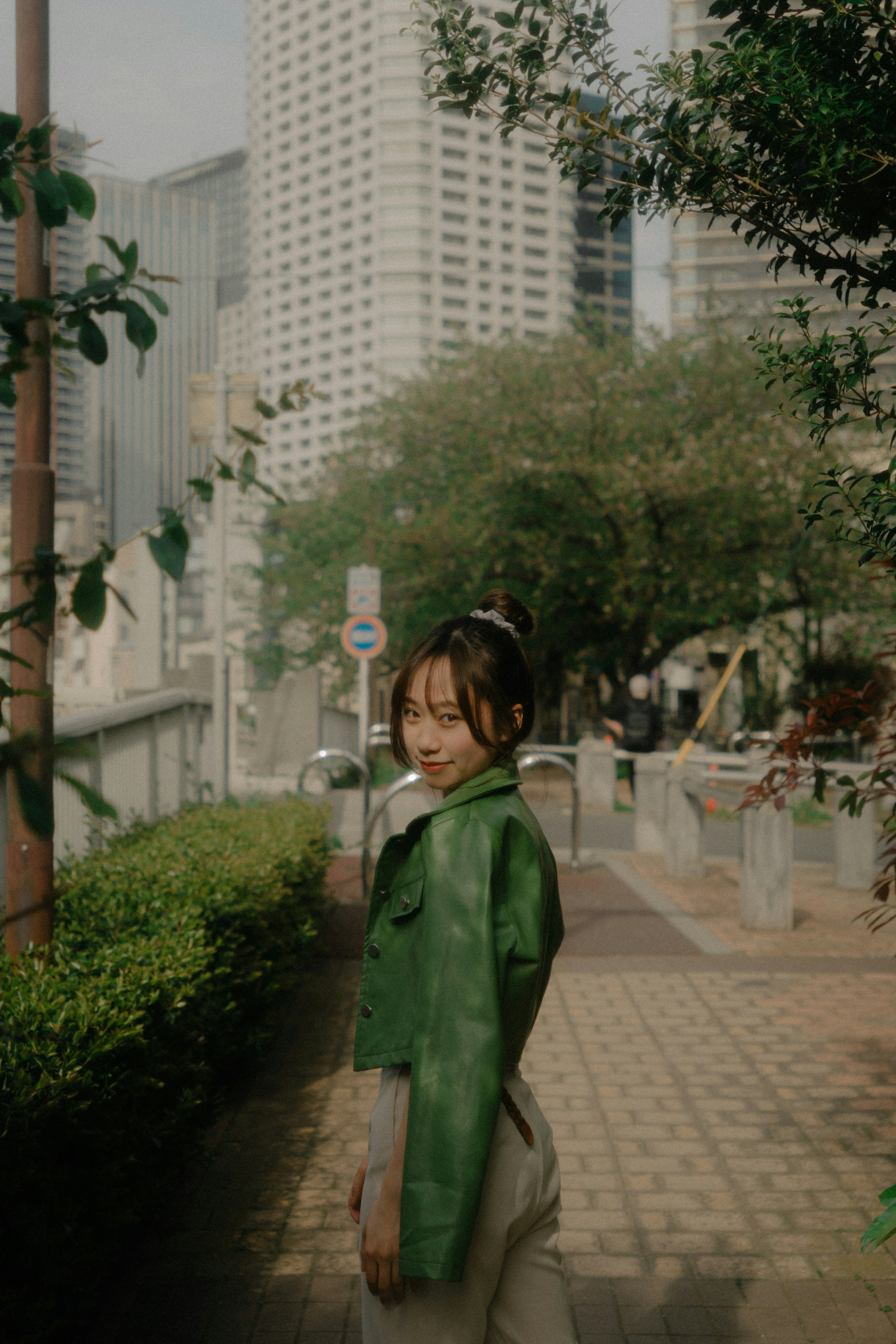 A woman in a green jacket turns back on an urban sidewalk with skyscrapers in the background