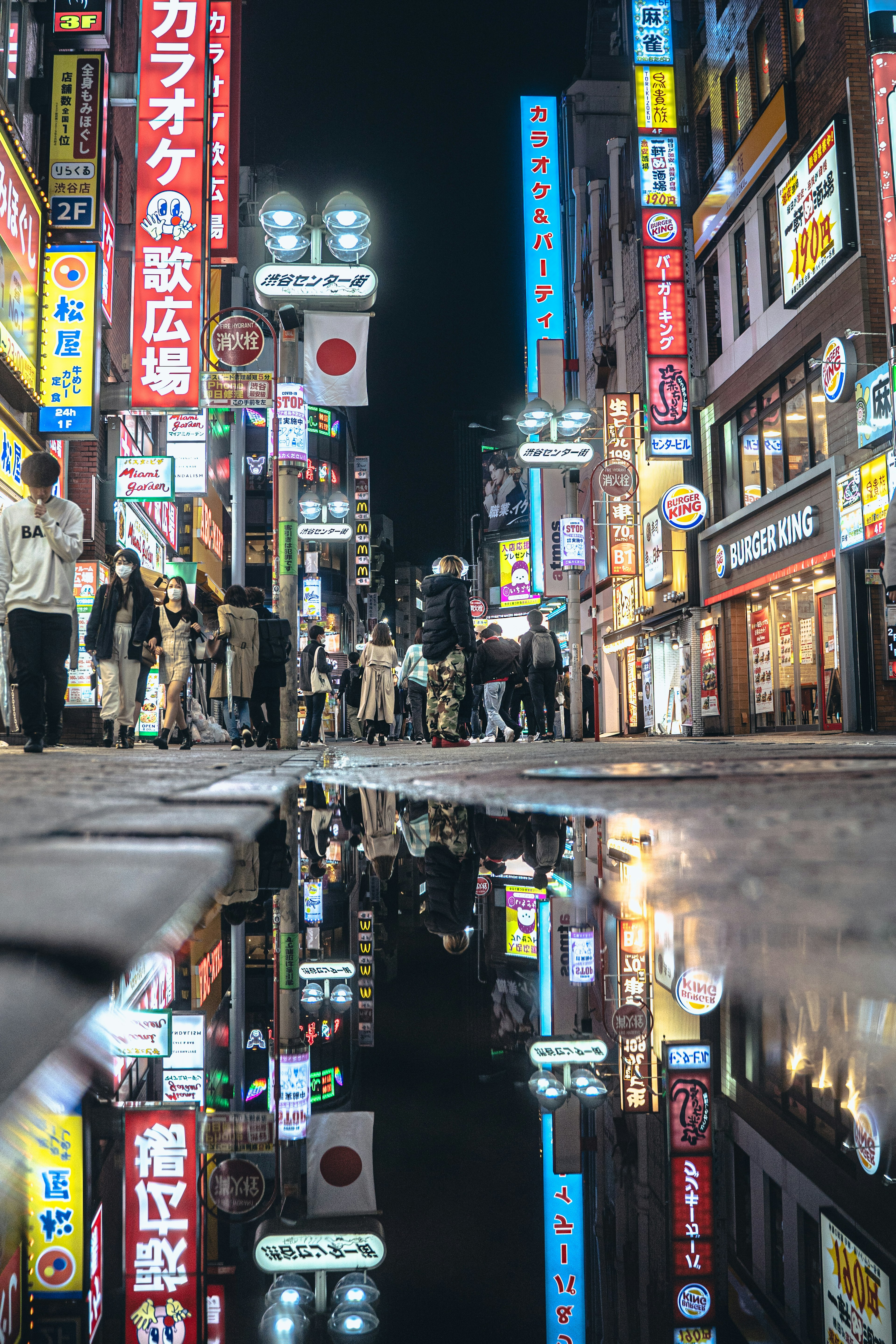 Colorful neon signs and people walking in a nighttime cityscape reflected in a puddle