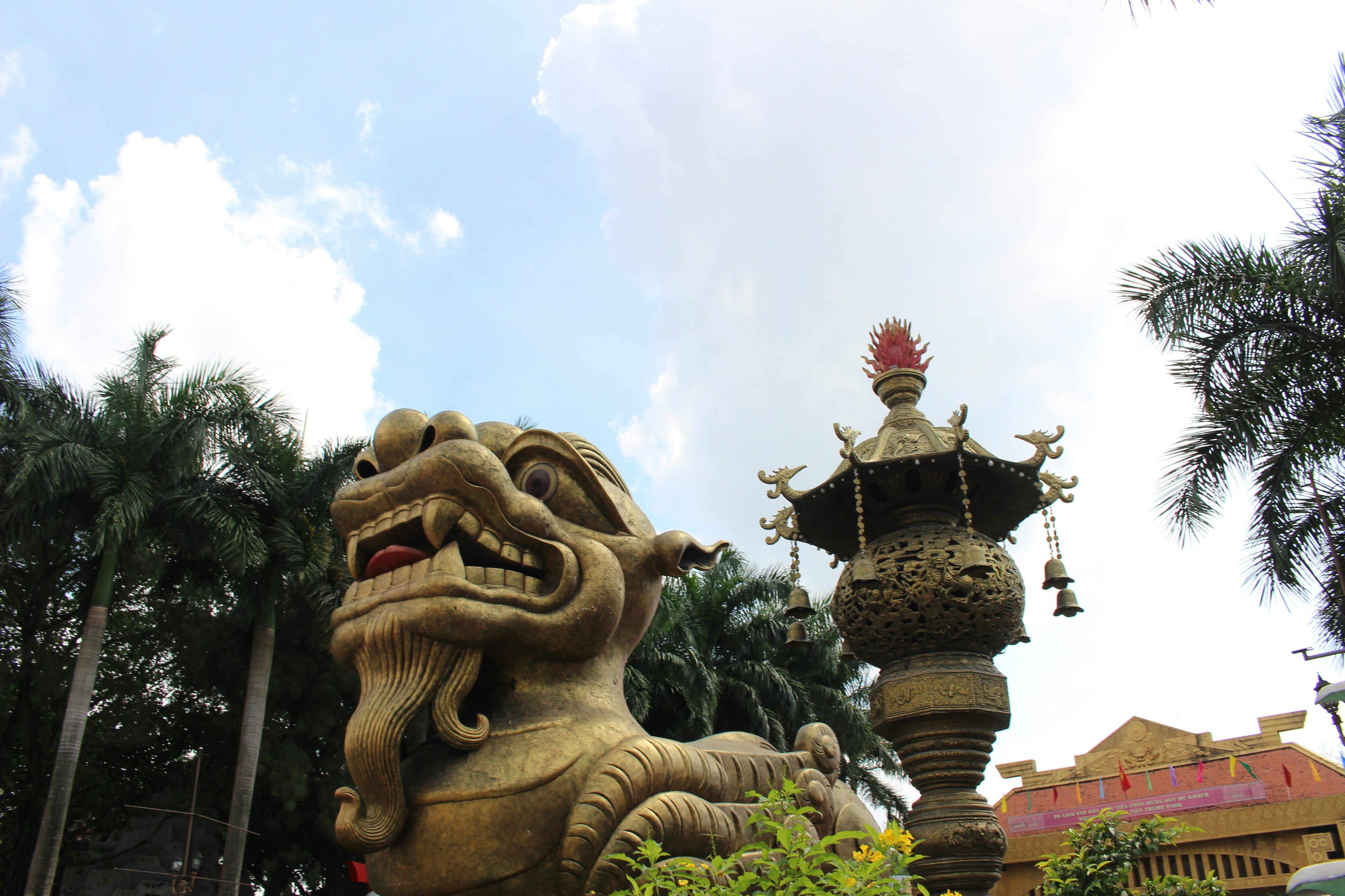 Gran estatua de león junto a una linterna decorativa en un jardín frondoso bajo un cielo azul y nubes blancas