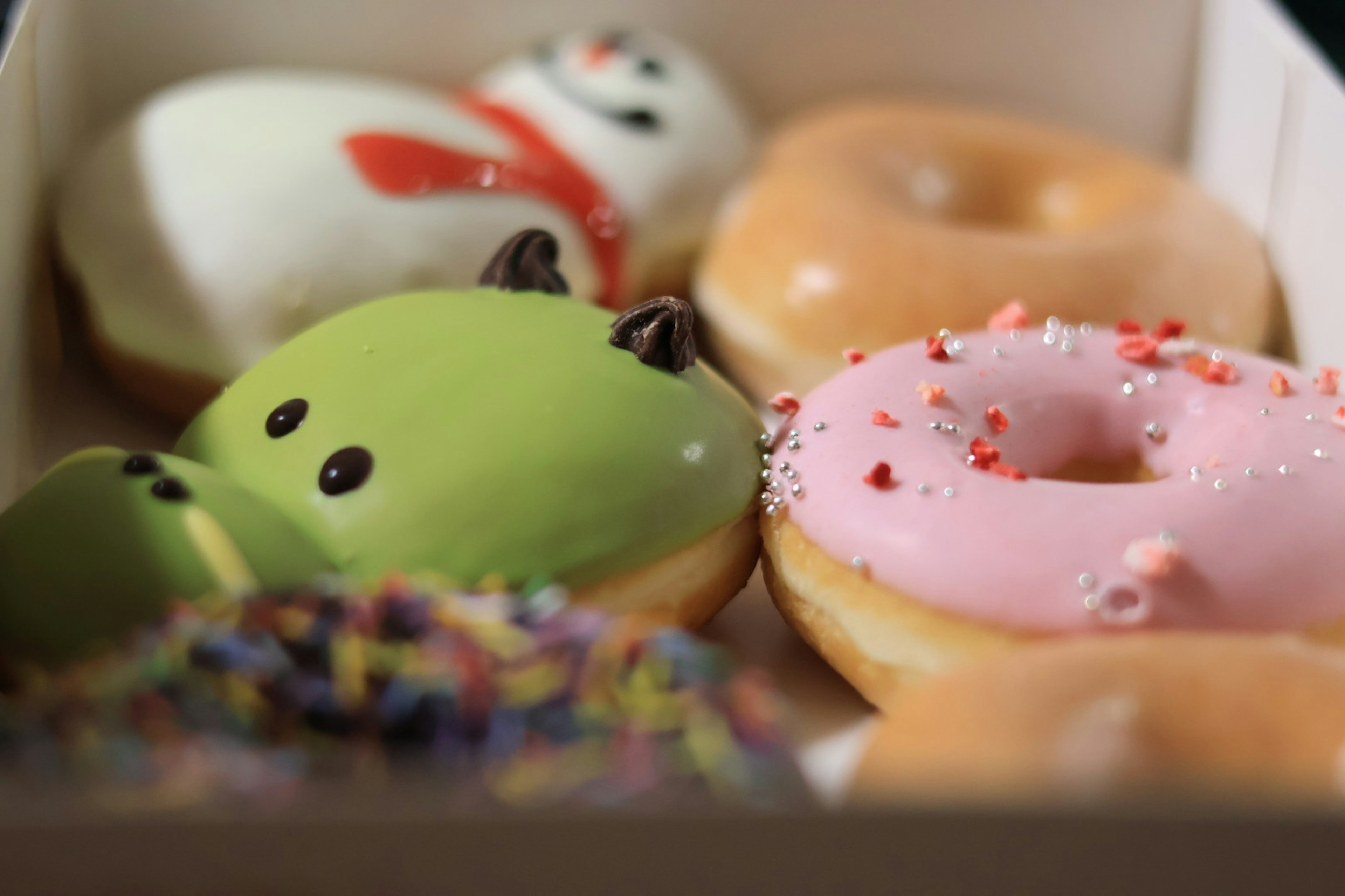 Eine Schachtel mit bunten Donuts, darunter ein Schneemann, eine grüne Figur und ein rosa Donut