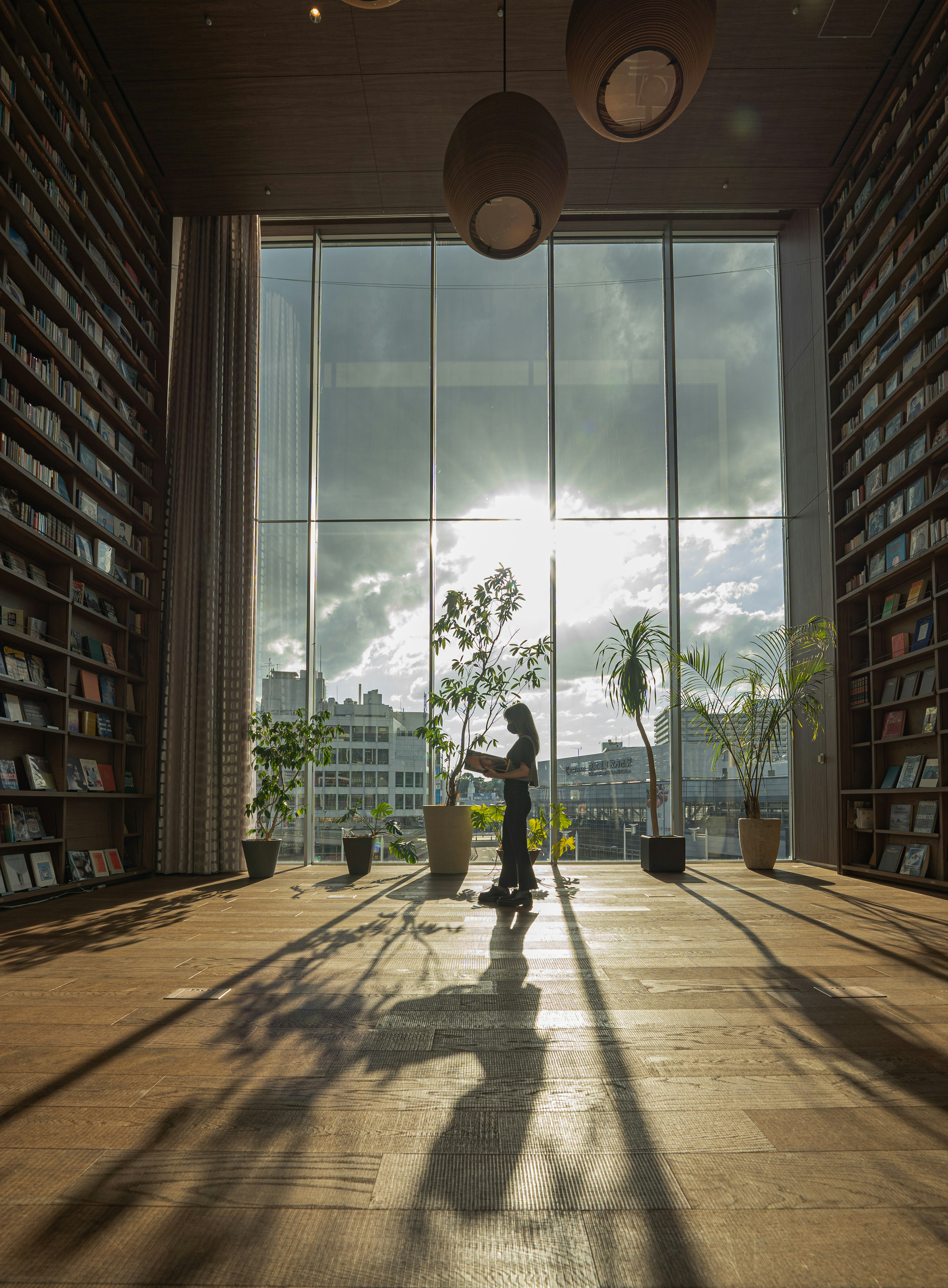 Interno di una biblioteca con grandi finestre che lasciano entrare la luce Una persona in piedi con un libro la cui silhouette proietta ombre sul pavimento