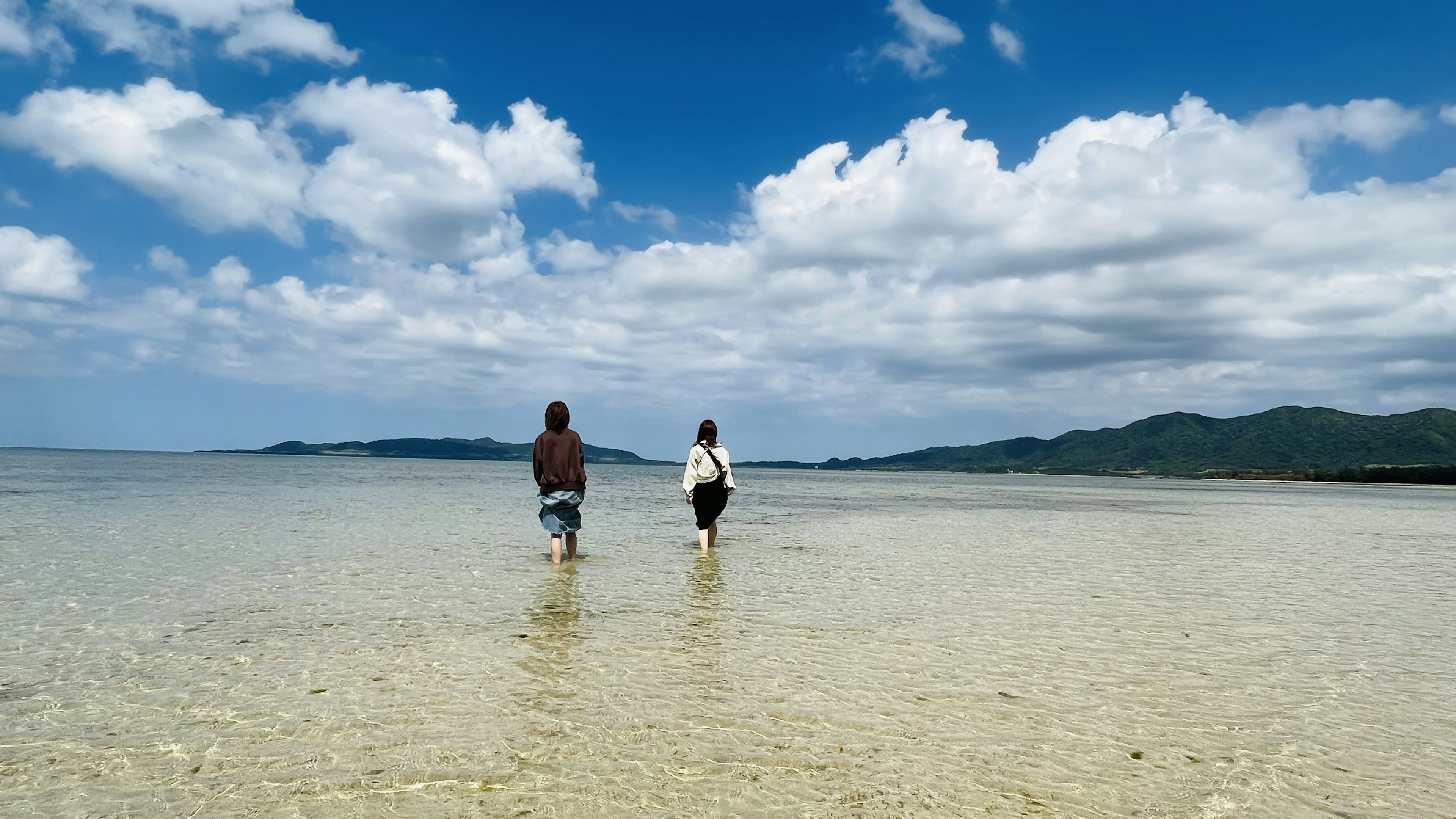 Dos personas caminando por la orilla bajo un cielo azul con nubes blancas agua clara