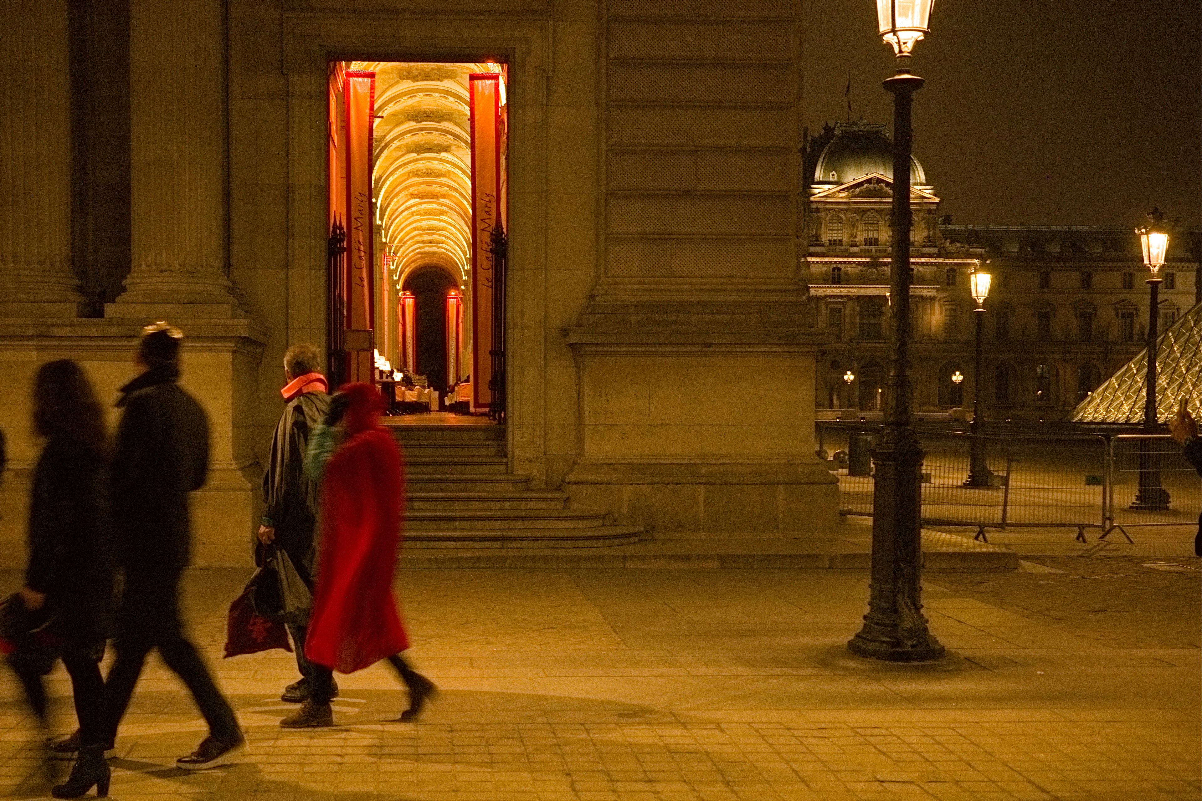 Personas caminando frente al Museo del Louvre por la noche una mujer con abrigo rojo destaca