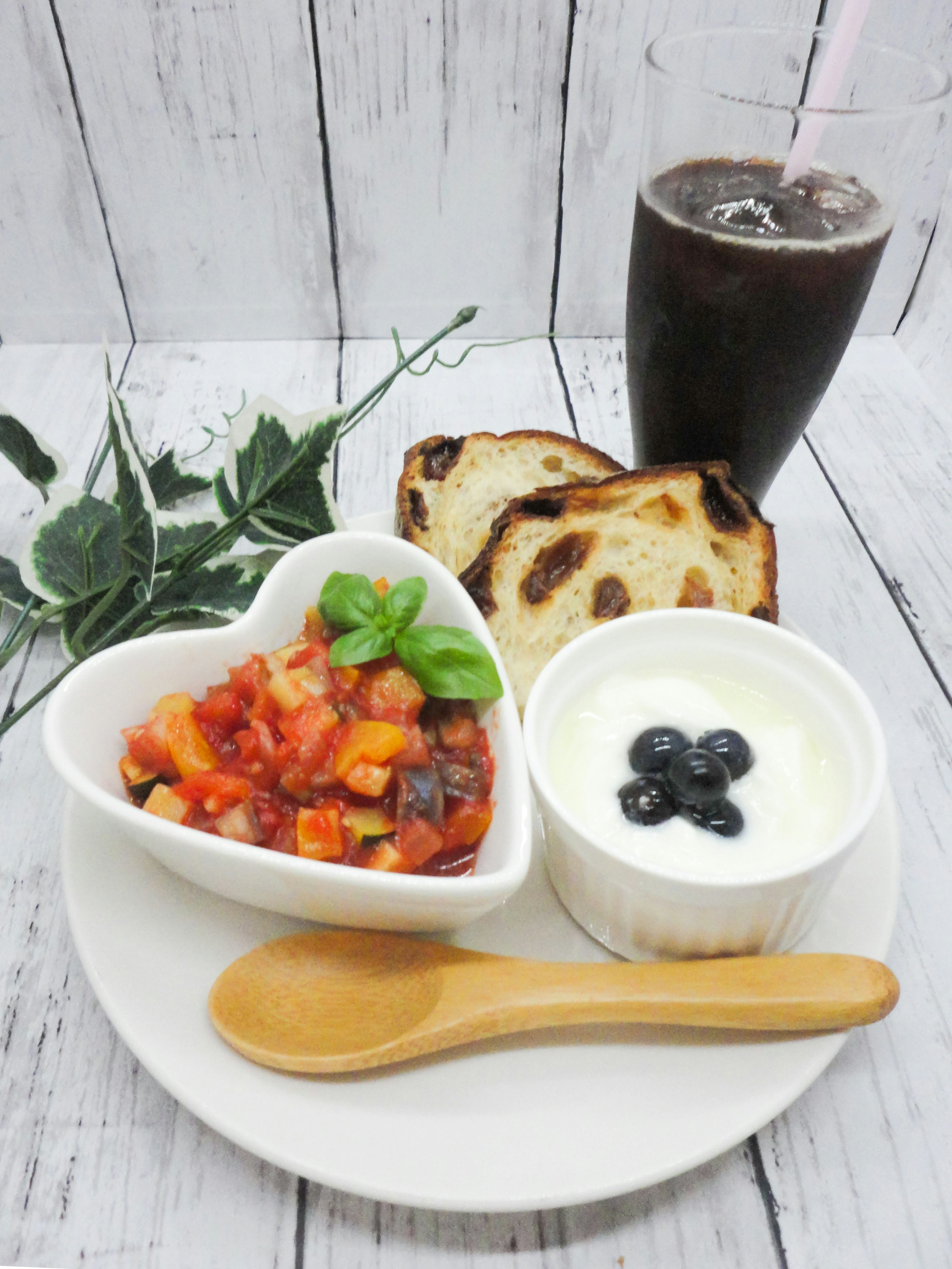 A heart-shaped dish with vegetable salad and yogurt topped with blueberries alongside toasted bread and a drink