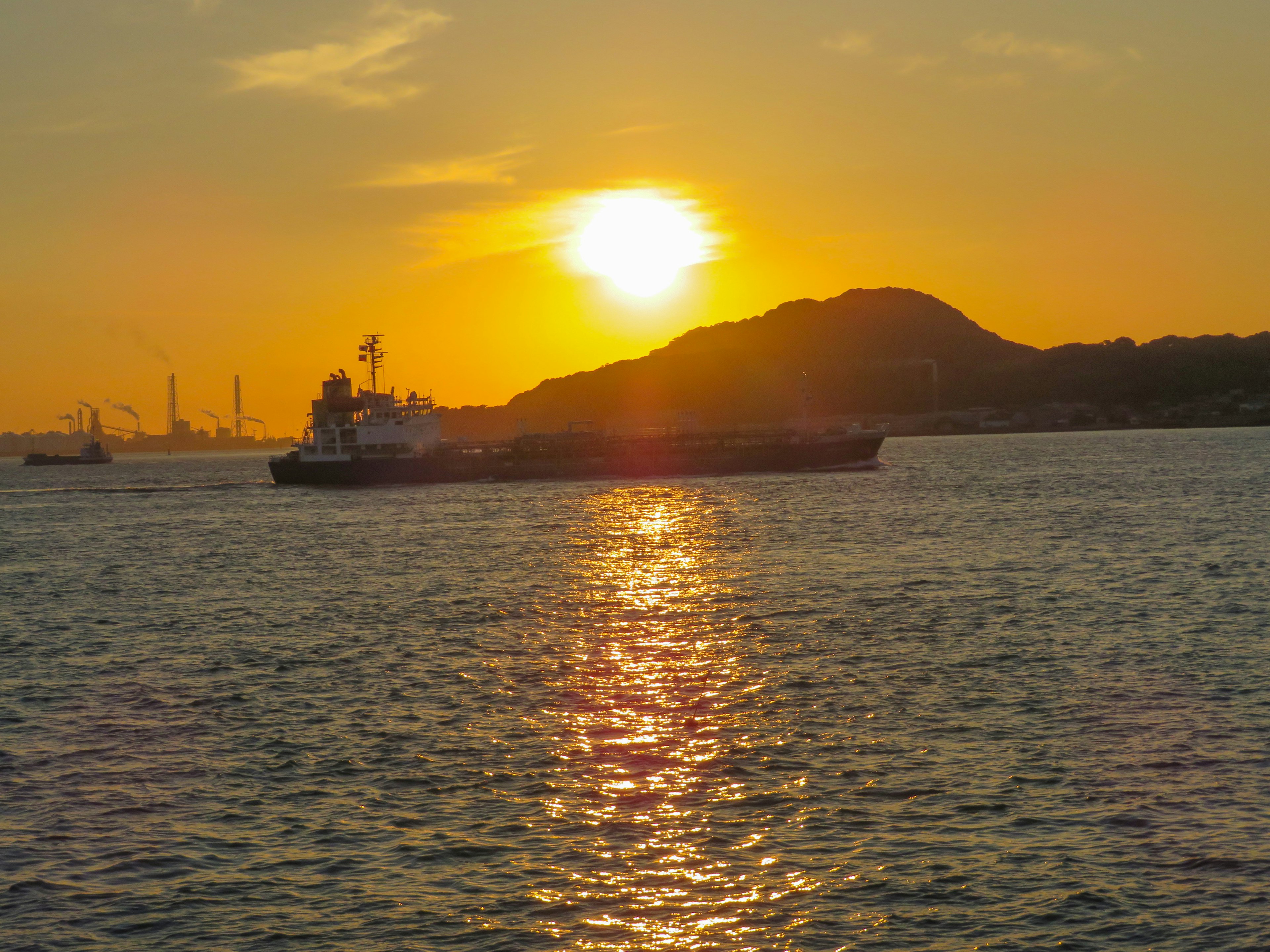 Sonnenuntergang über dem Ozean mit einem Schiff, das über das Wasser gleitet Berge und Fabriken im Hintergrund