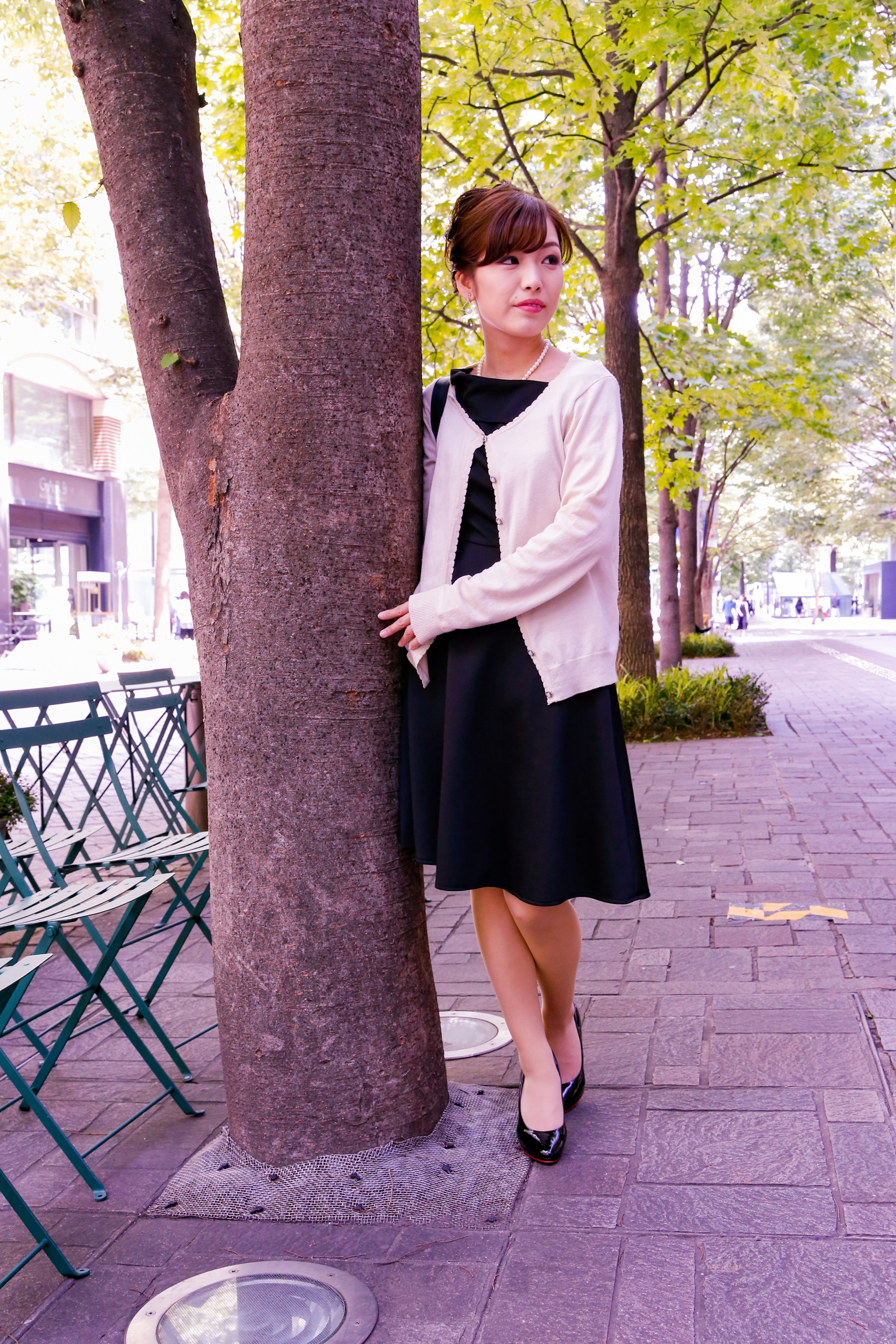 Mujer de pie junto a un árbol en un parque con un vestido negro y un cárdigan rosa en un entorno otoñal