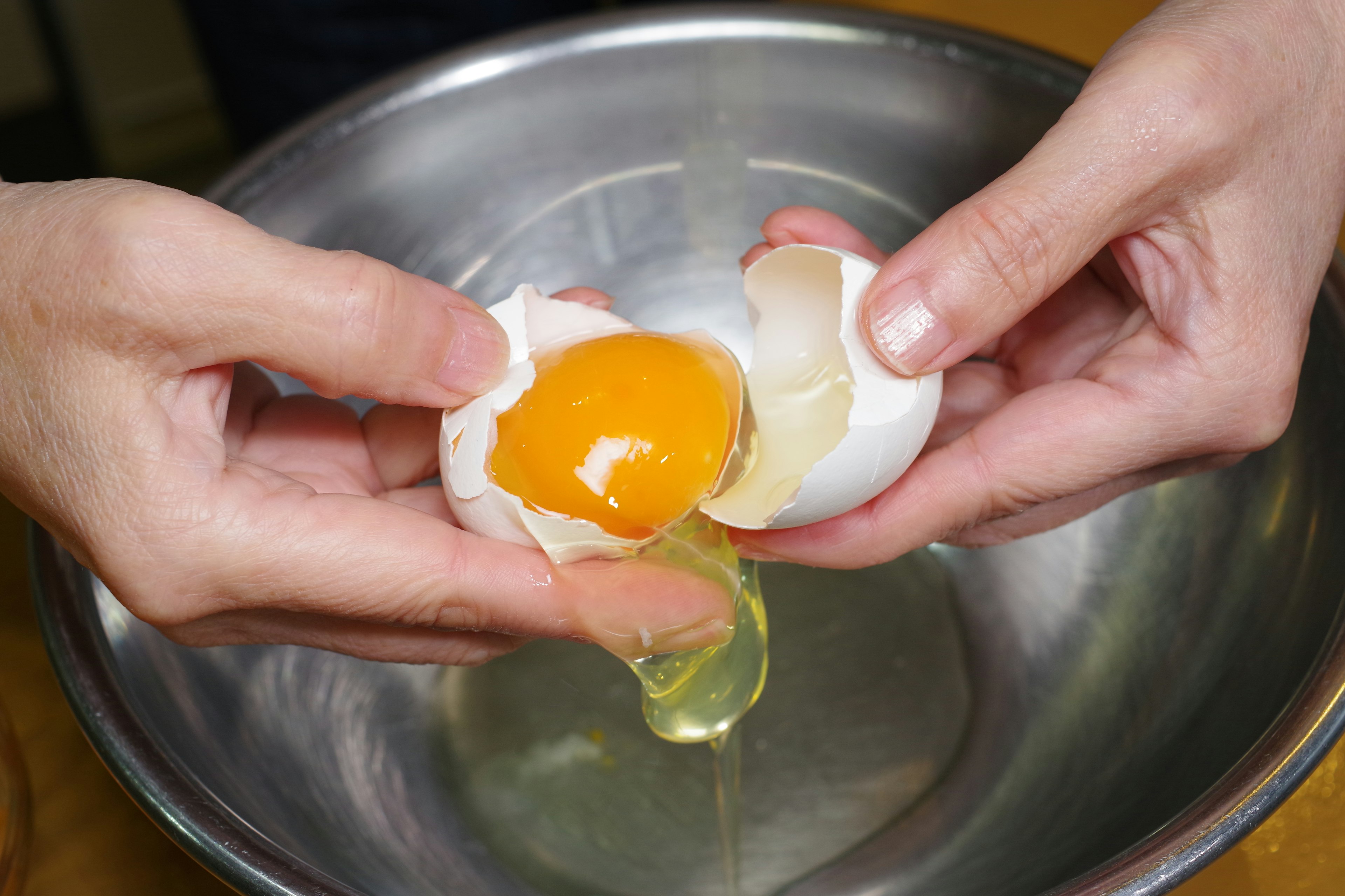 Close-up tangan memecahkan telur dengan kuning dan putih telur jatuh ke dalam mangkuk