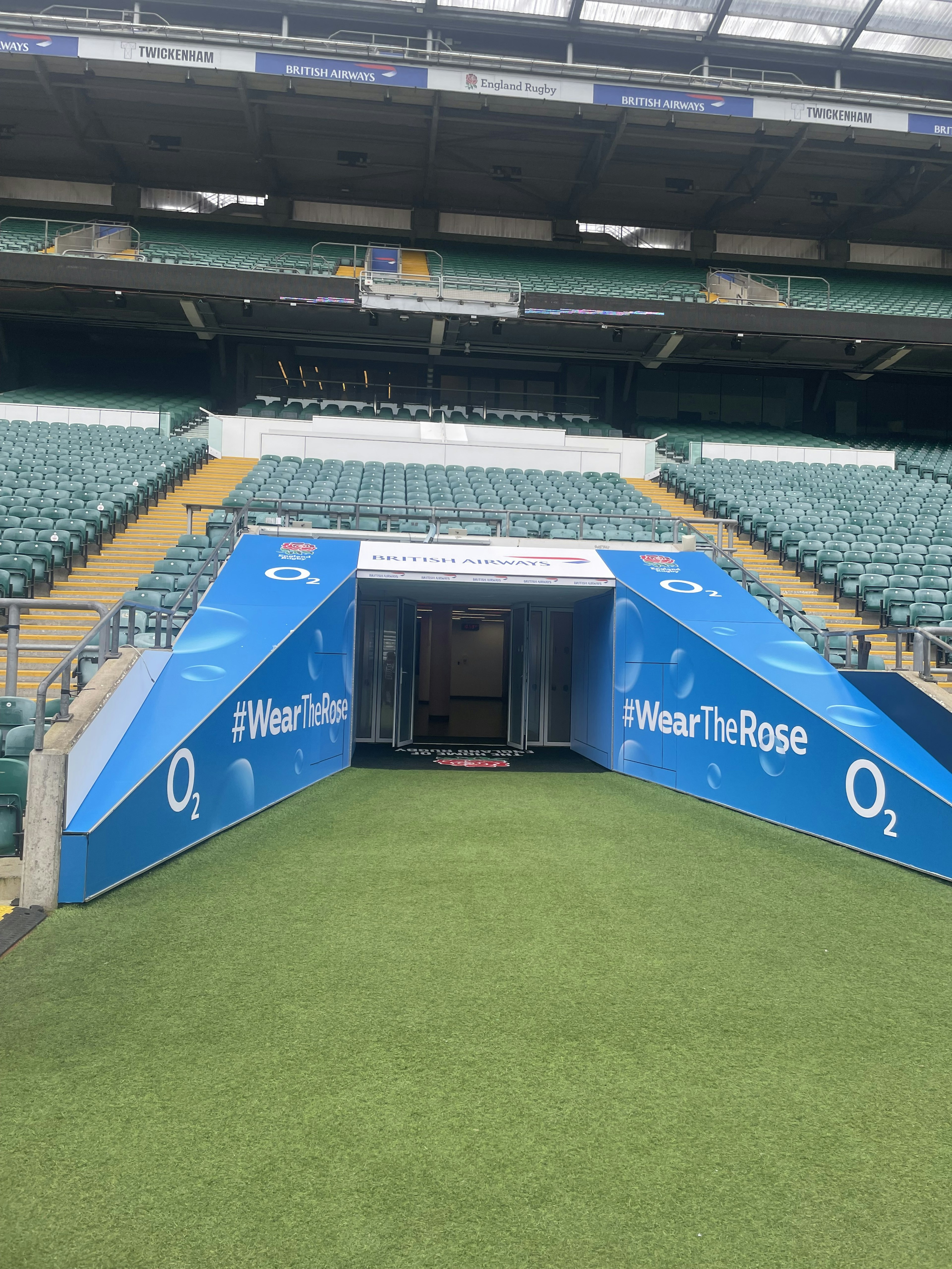 Pintu masuk terowongan stadion dengan spanduk biru dan rumput