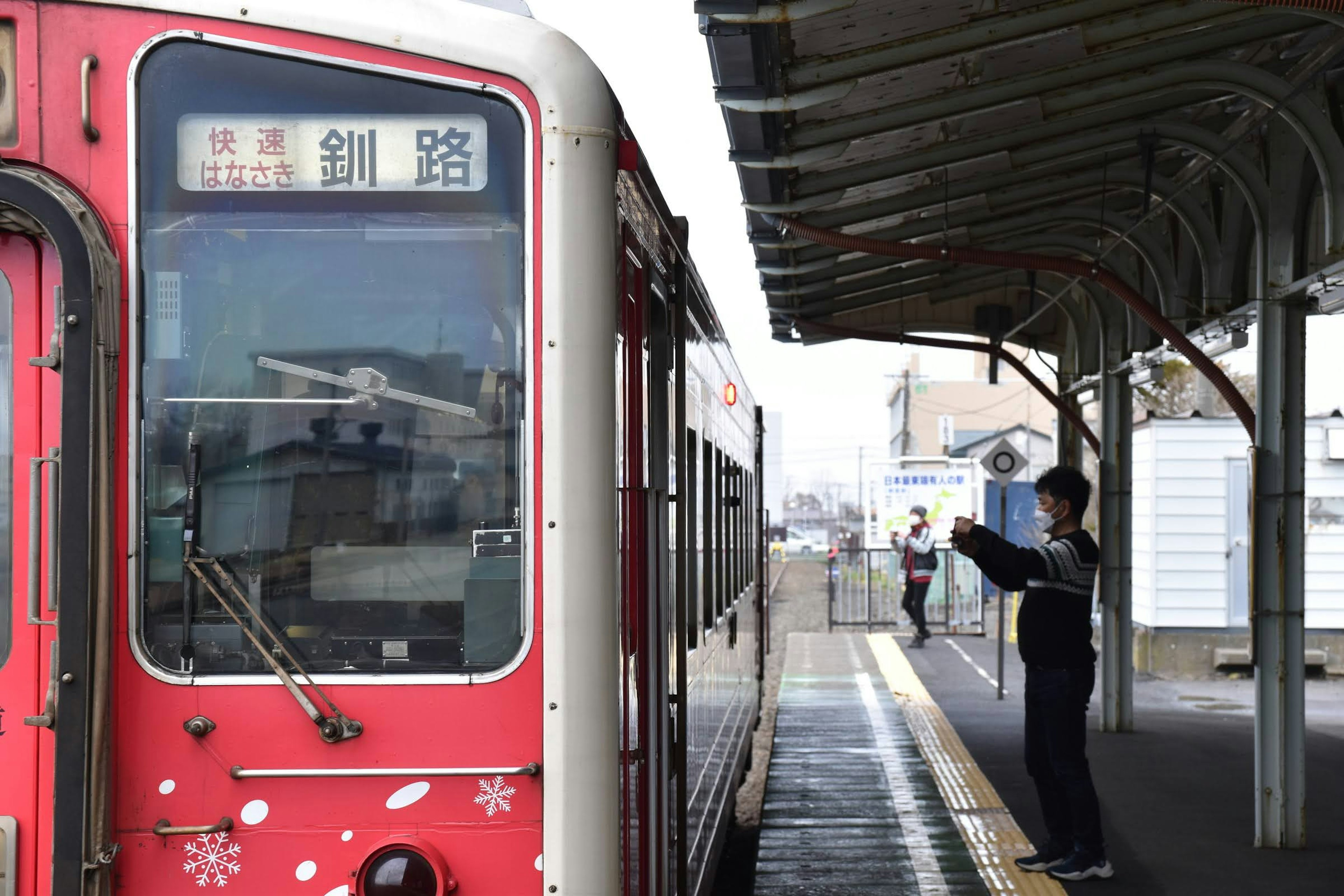 赤い電車が駅に停車し、ホームに立つ人がいる風景