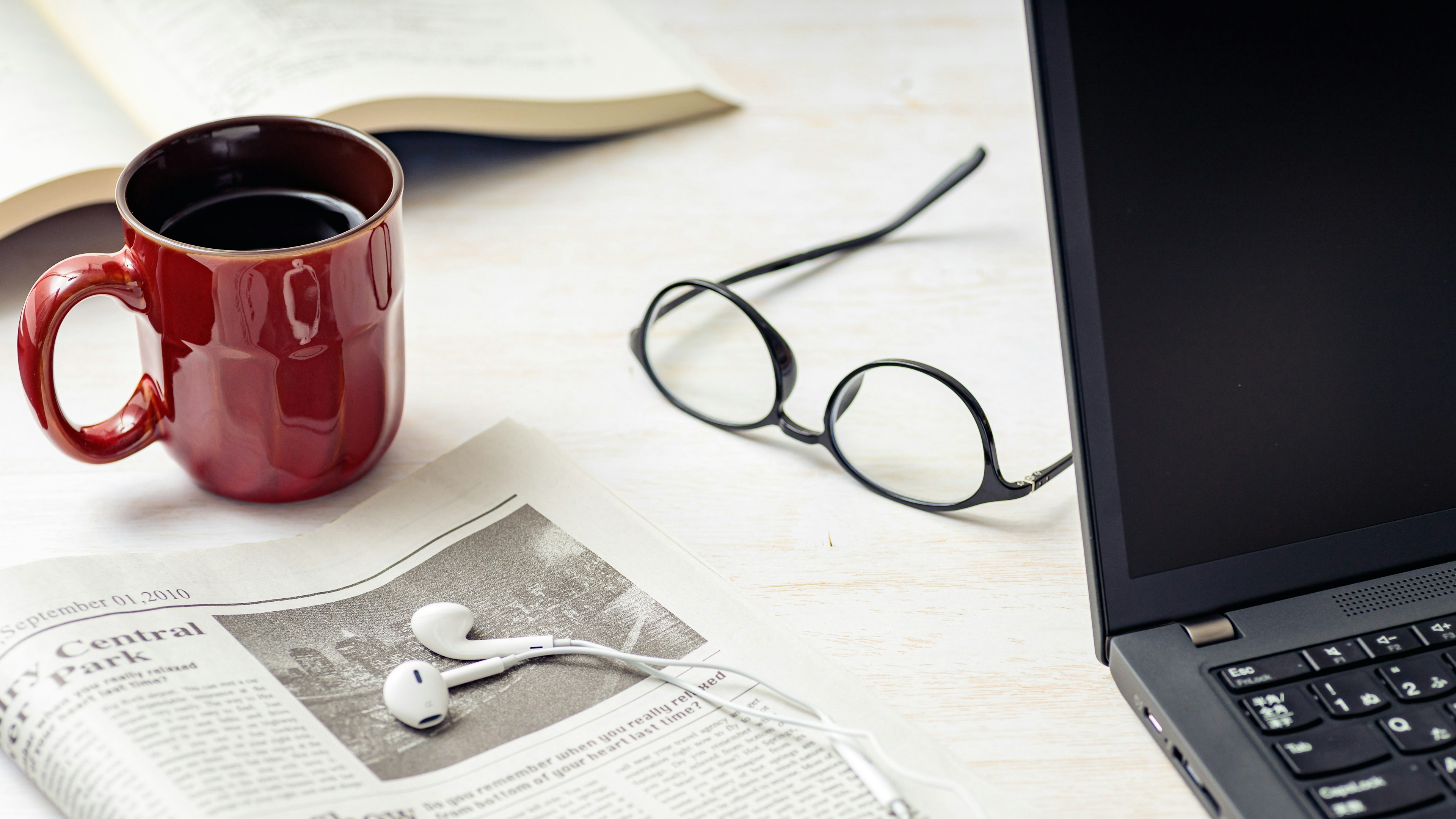 A red coffee mug and newspaper on a table with a laptop and glasses