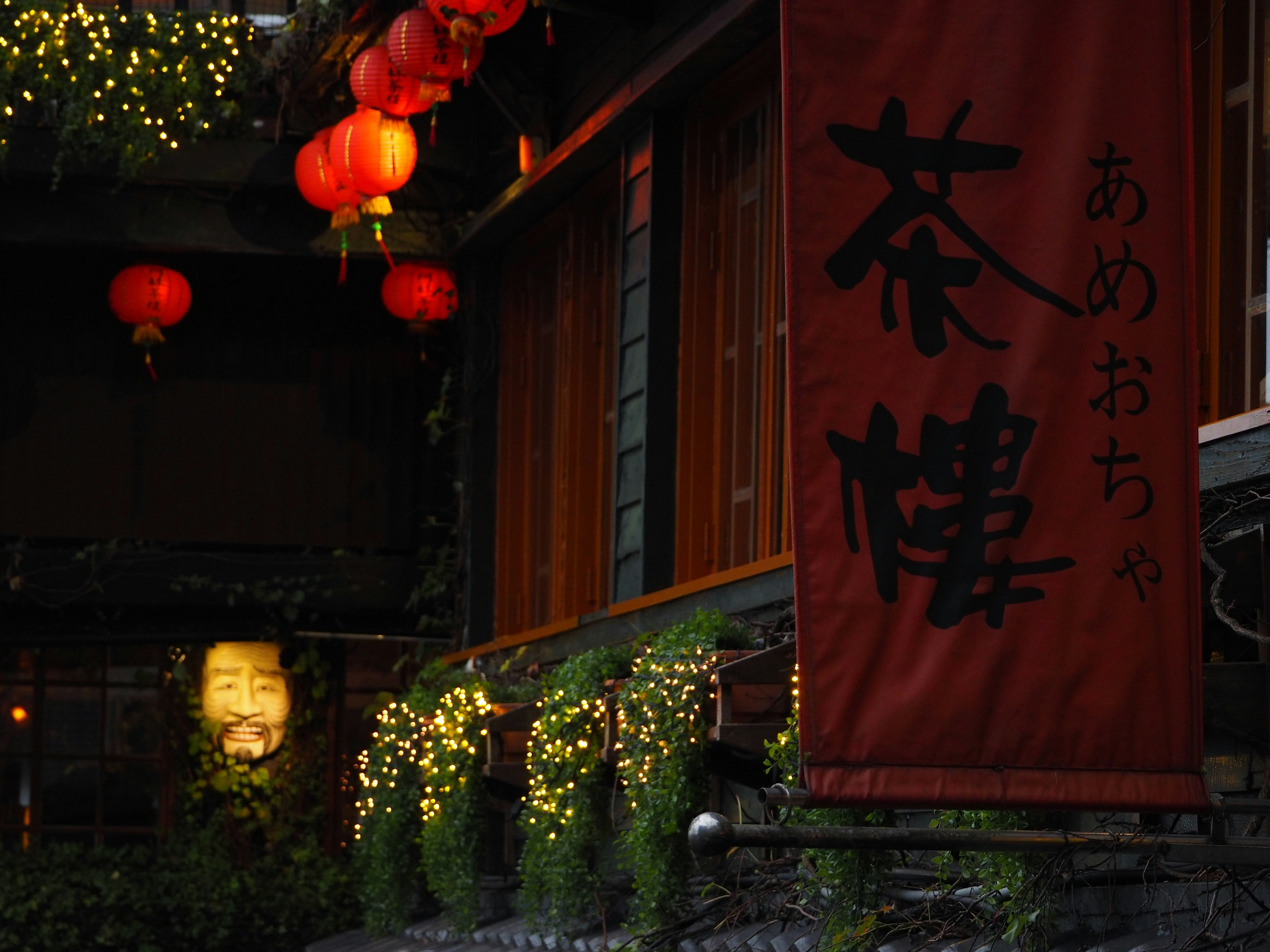 Vista nocturna de una casa de té con faroles rojos y decoraciones de plantas verdes