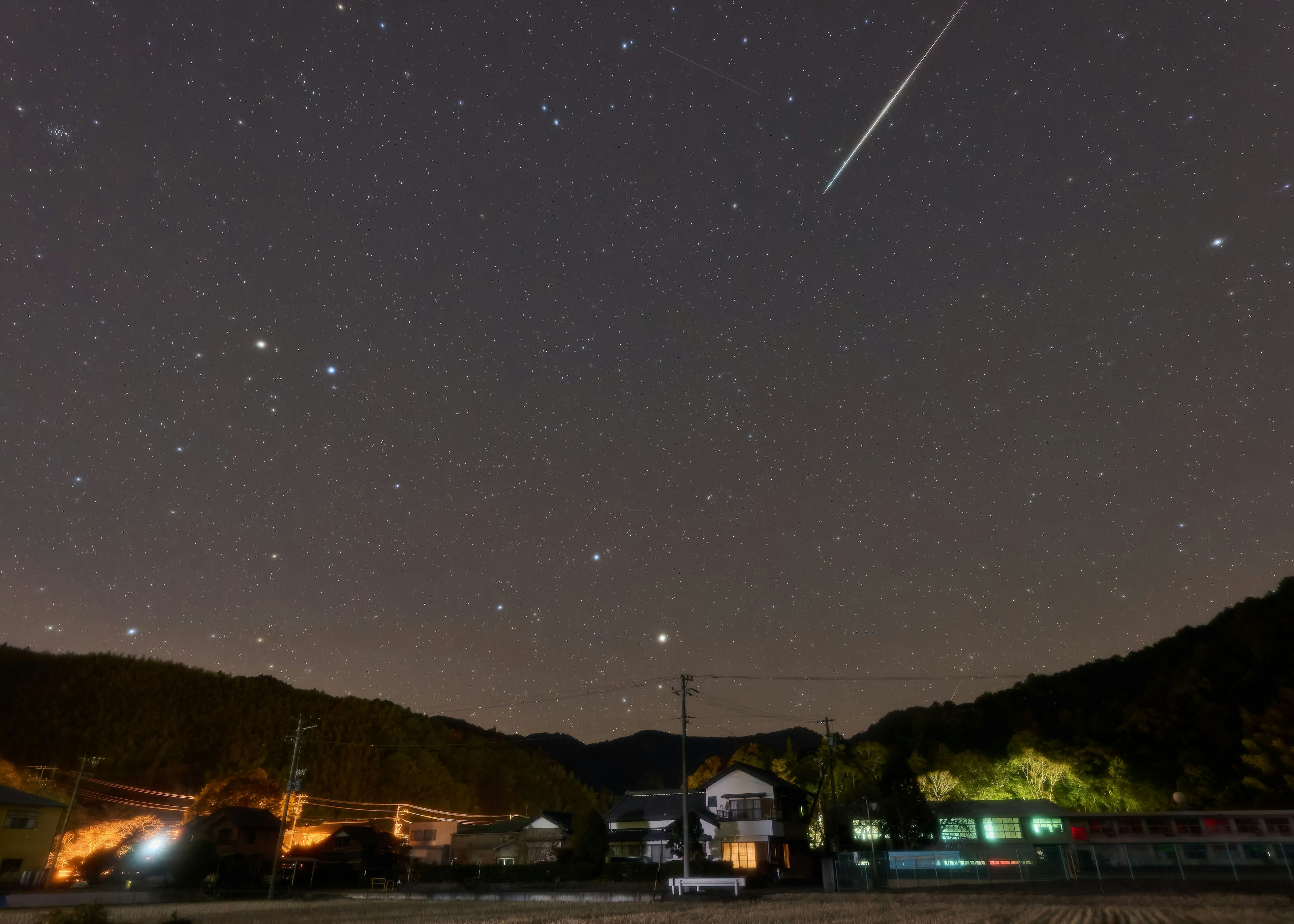 Night landscape featuring a starry sky and a meteor low buildings and mountains in the background