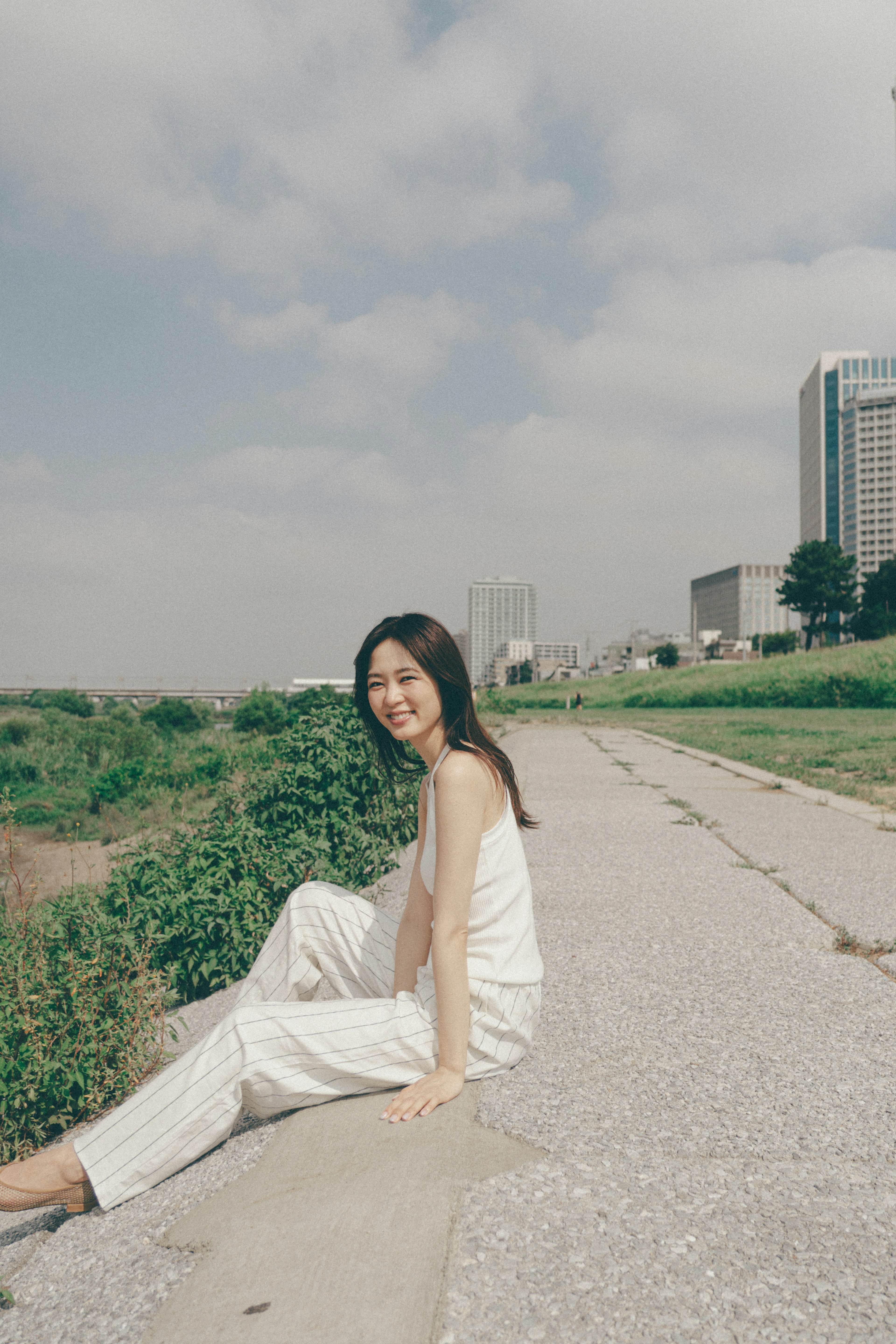 A woman in white clothing sitting on a pathway near a river under bright sunlight