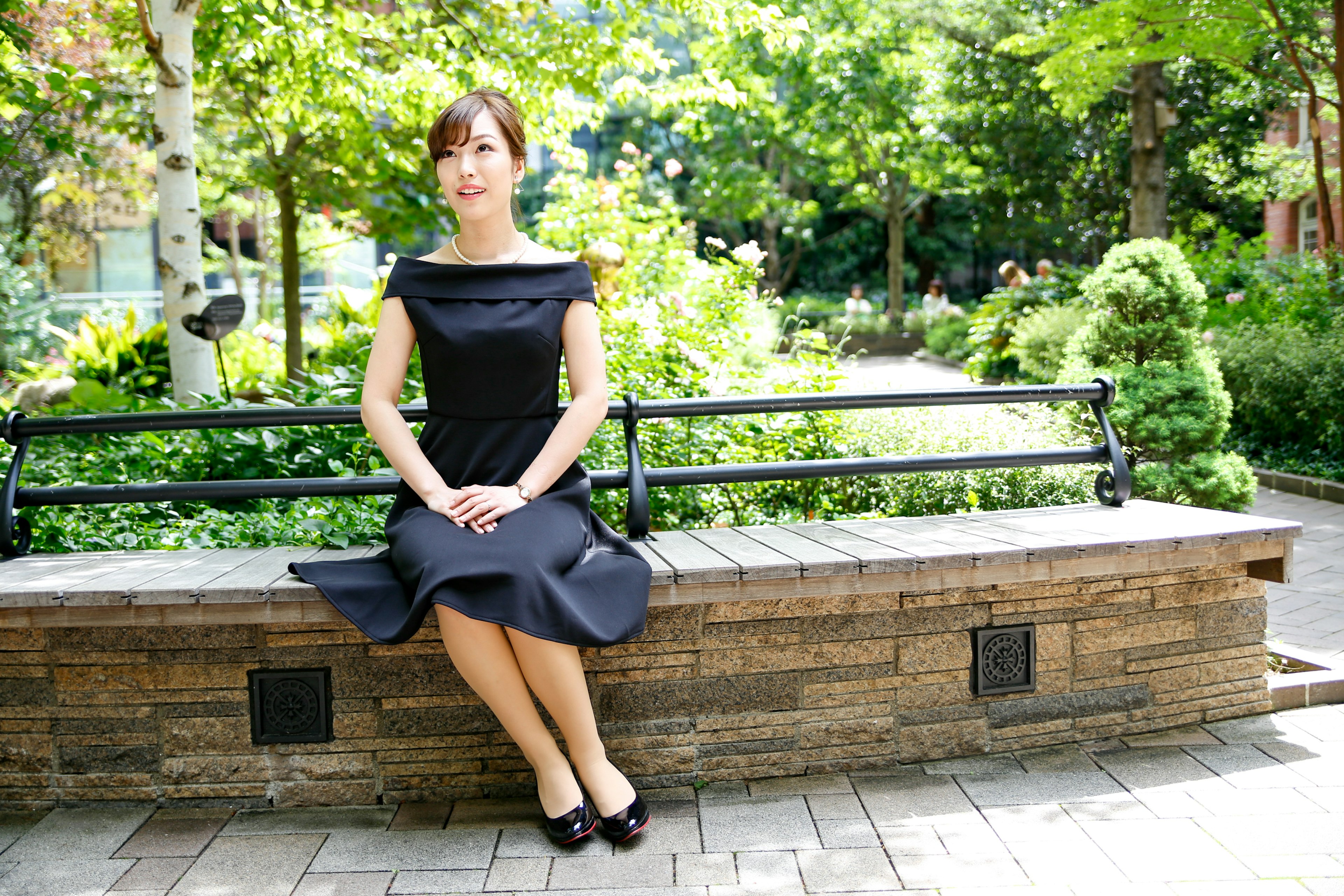 Femme en robe noire assise sur un banc de parc entourée de verdure