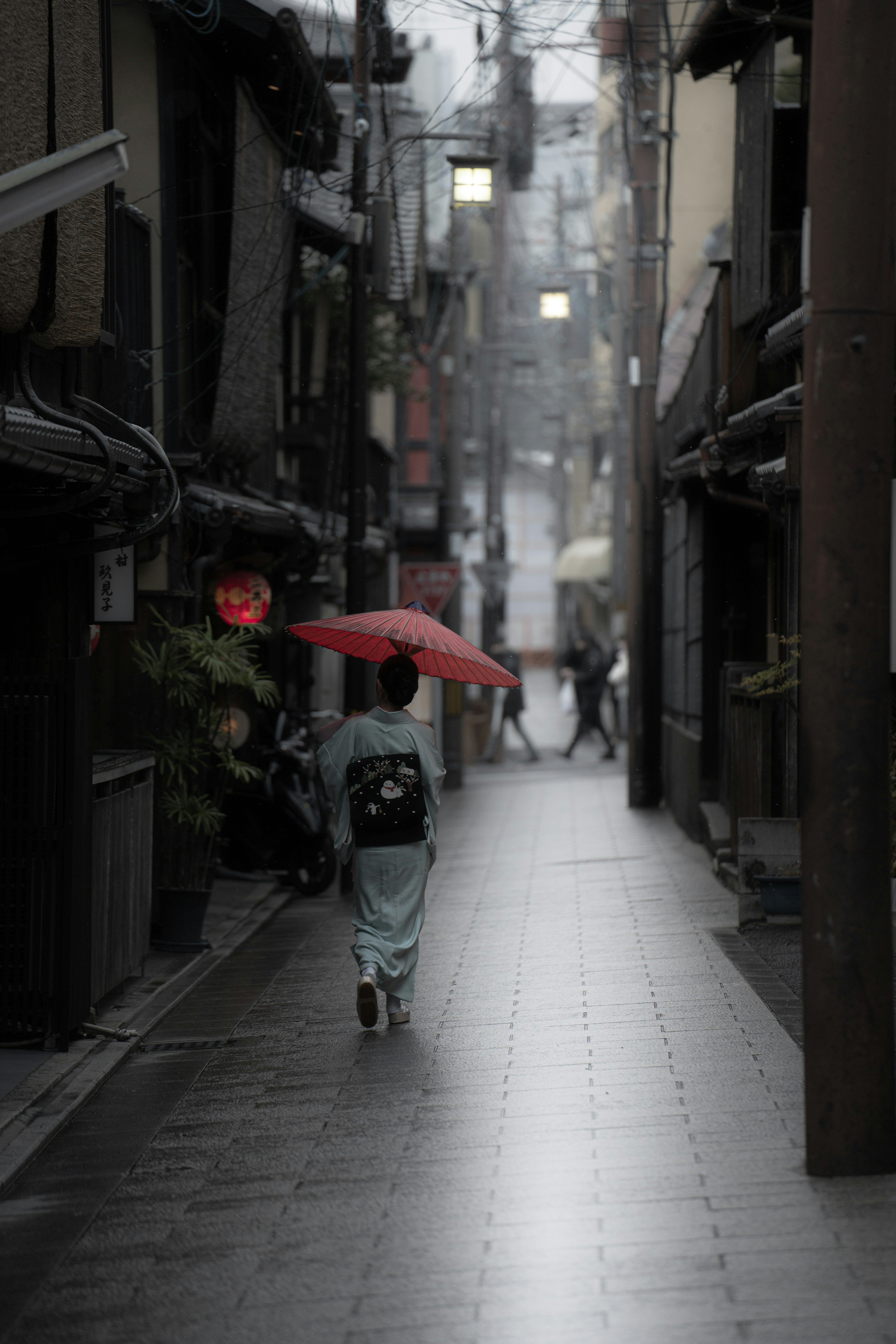 雨の中、赤い傘を持った人が狭い路地を歩いている風景