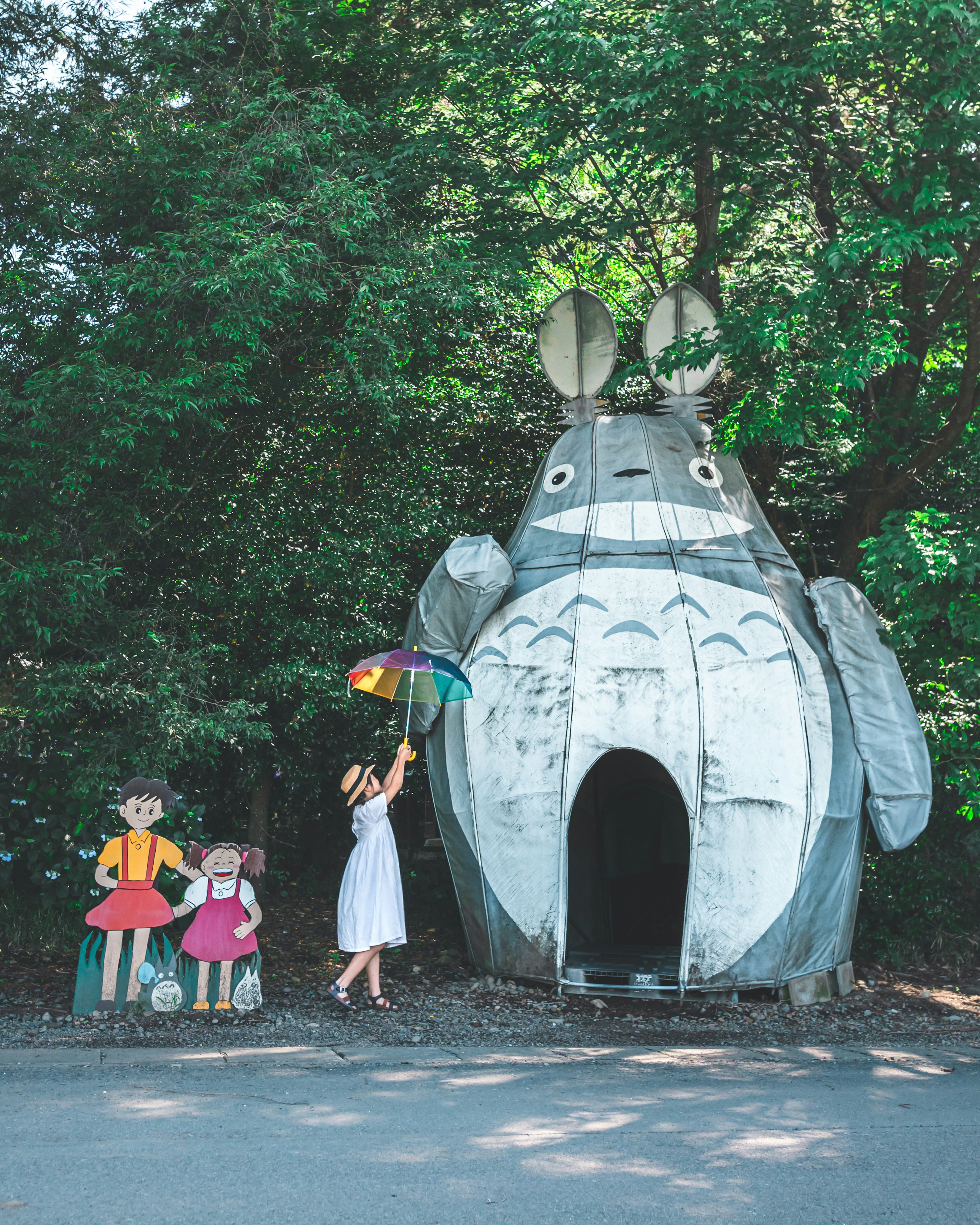 A girl holding an umbrella stands in front of a Totoro house with two characters nearby