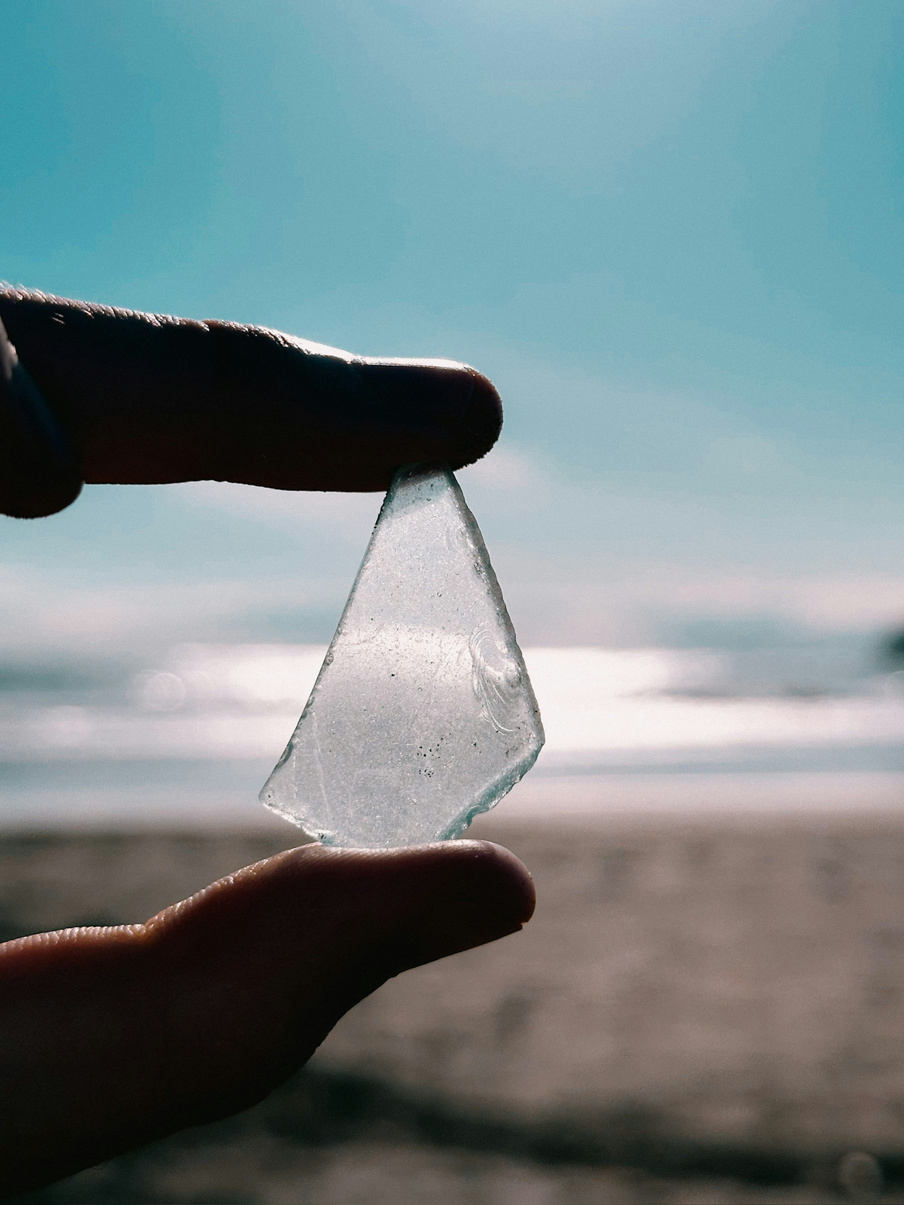 A hand holding a clear piece of glass against a beach background with a blue sky