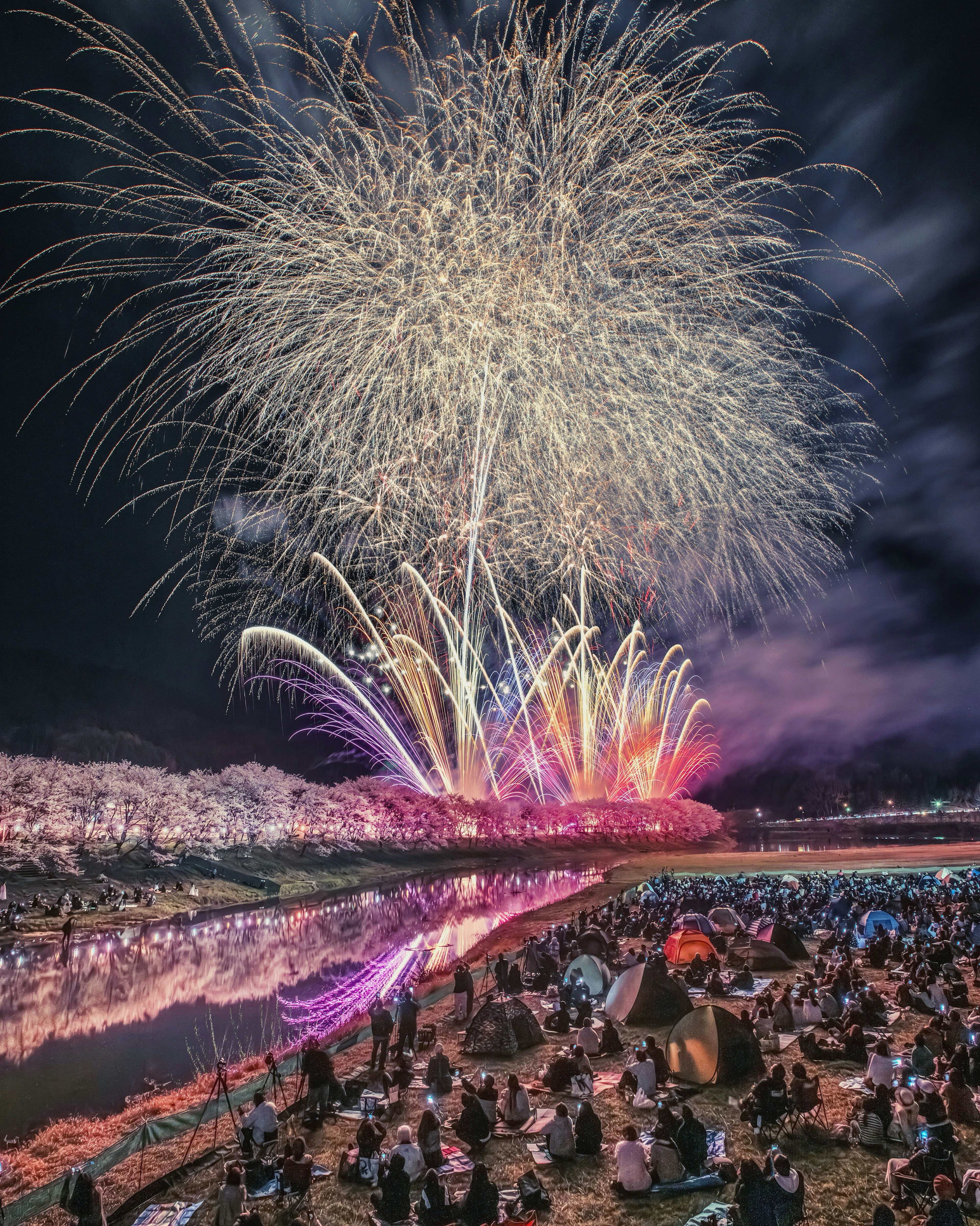 Spectacular fireworks display over a river with a large crowd gathered on the shore